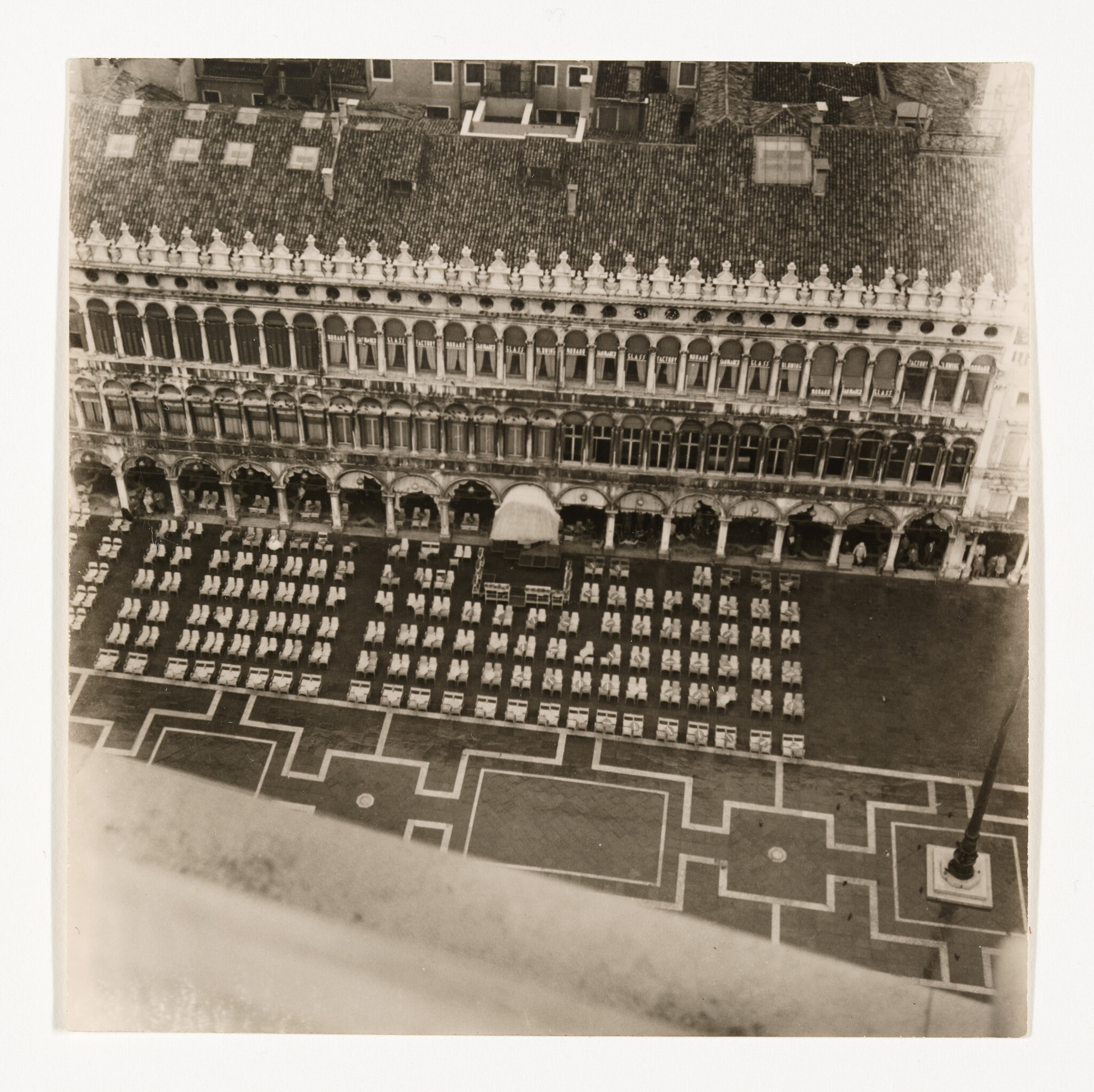 Aerial view of a long ornate arcade overlooking rows of neatly arranged chairs on a patterned plaza.