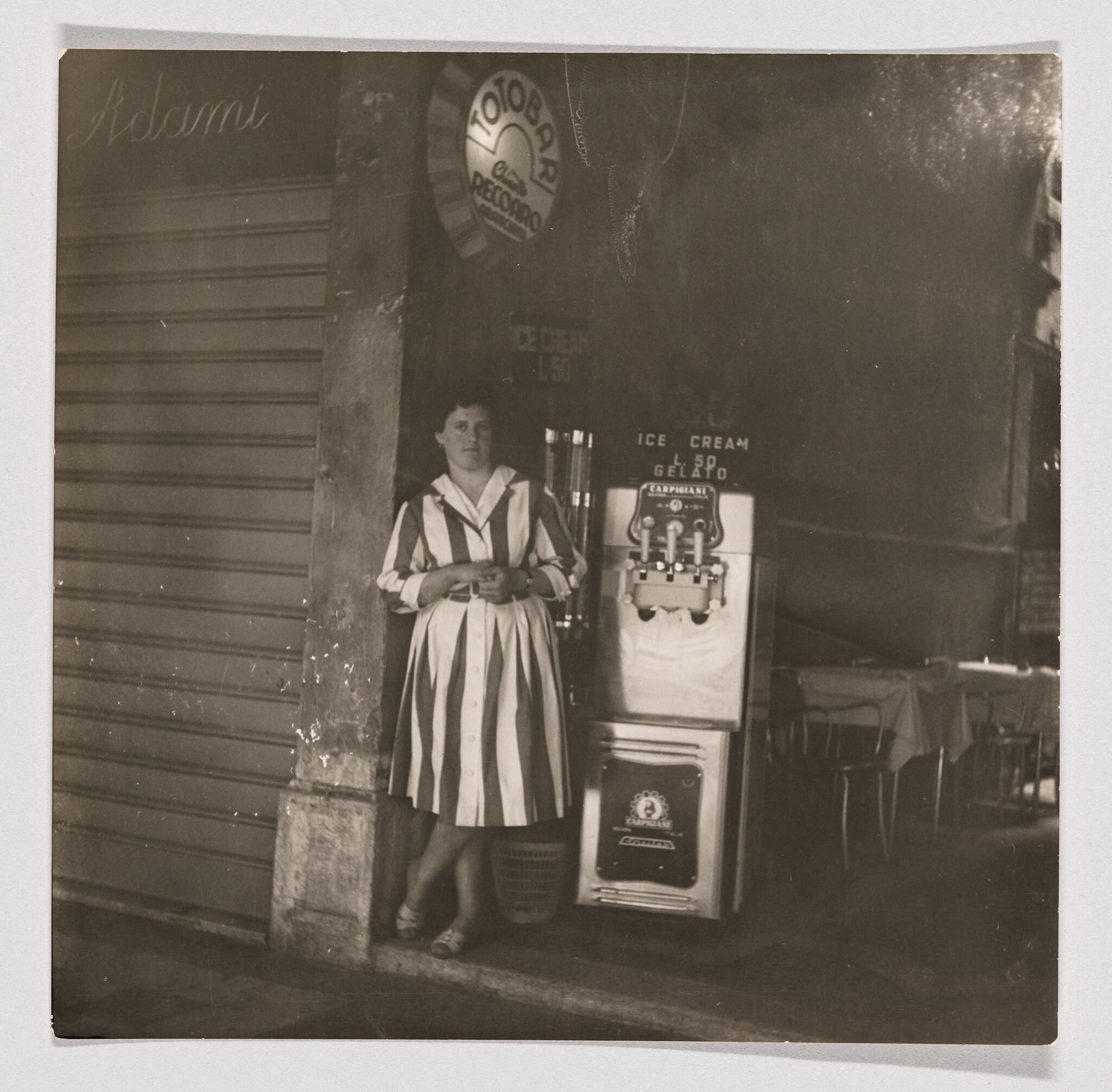 A woman in a striped dress stands beside an ice cream machine outside a café.