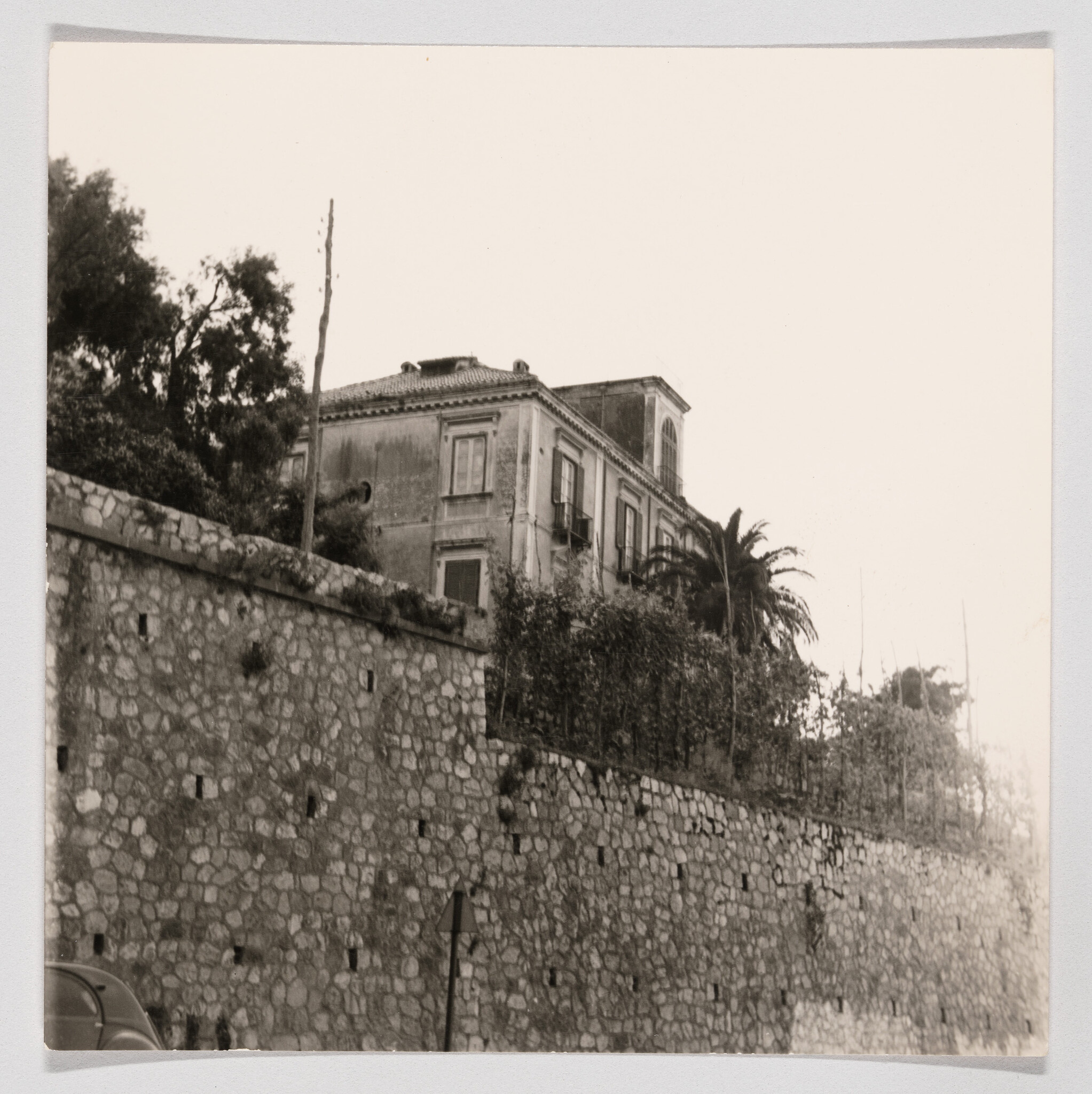 An old stucco house sits atop a tall stone retaining wall with a palm tree beside it.