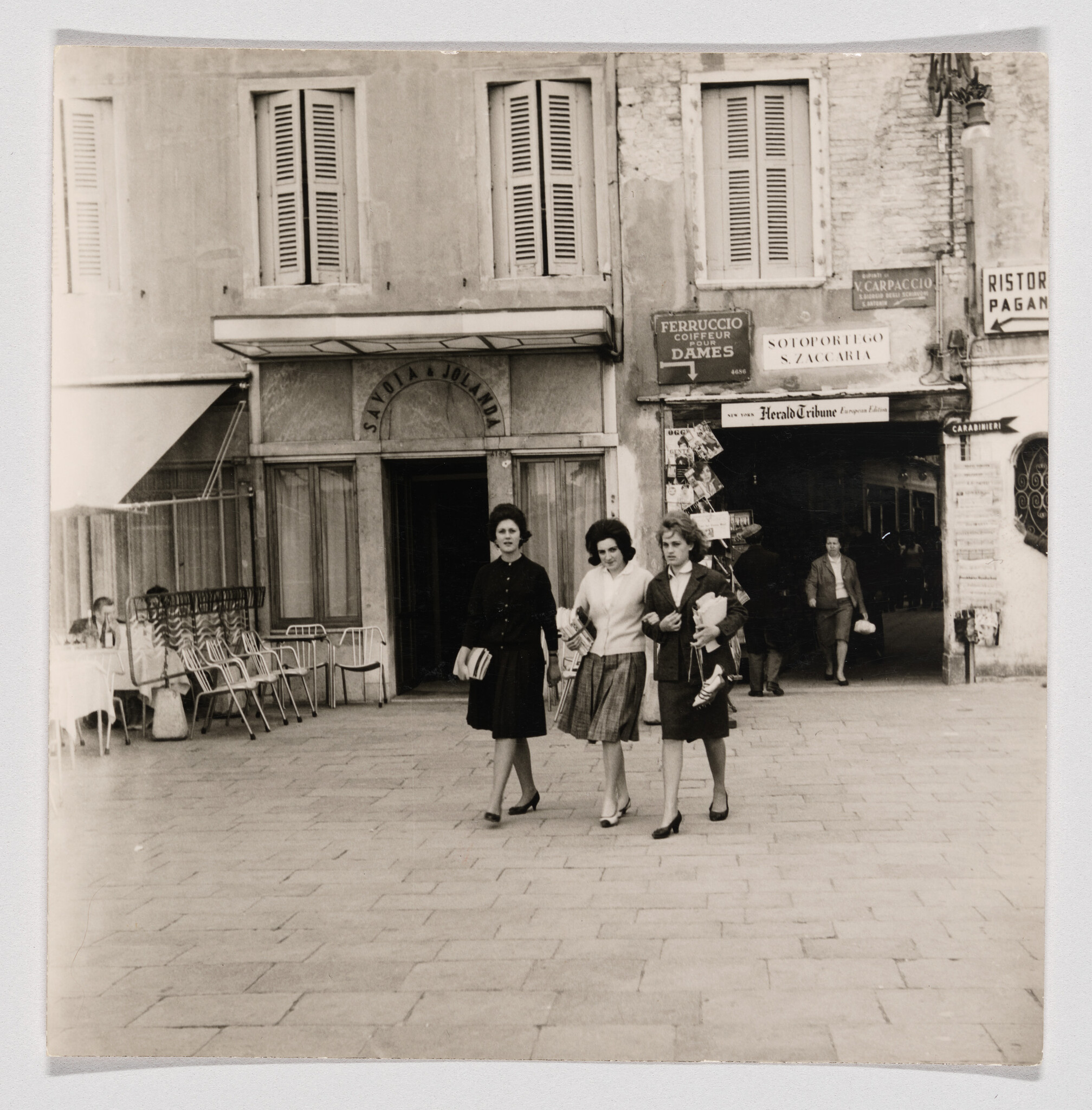 Three young women walk arm in arm across a town square past storefronts and a cafe.