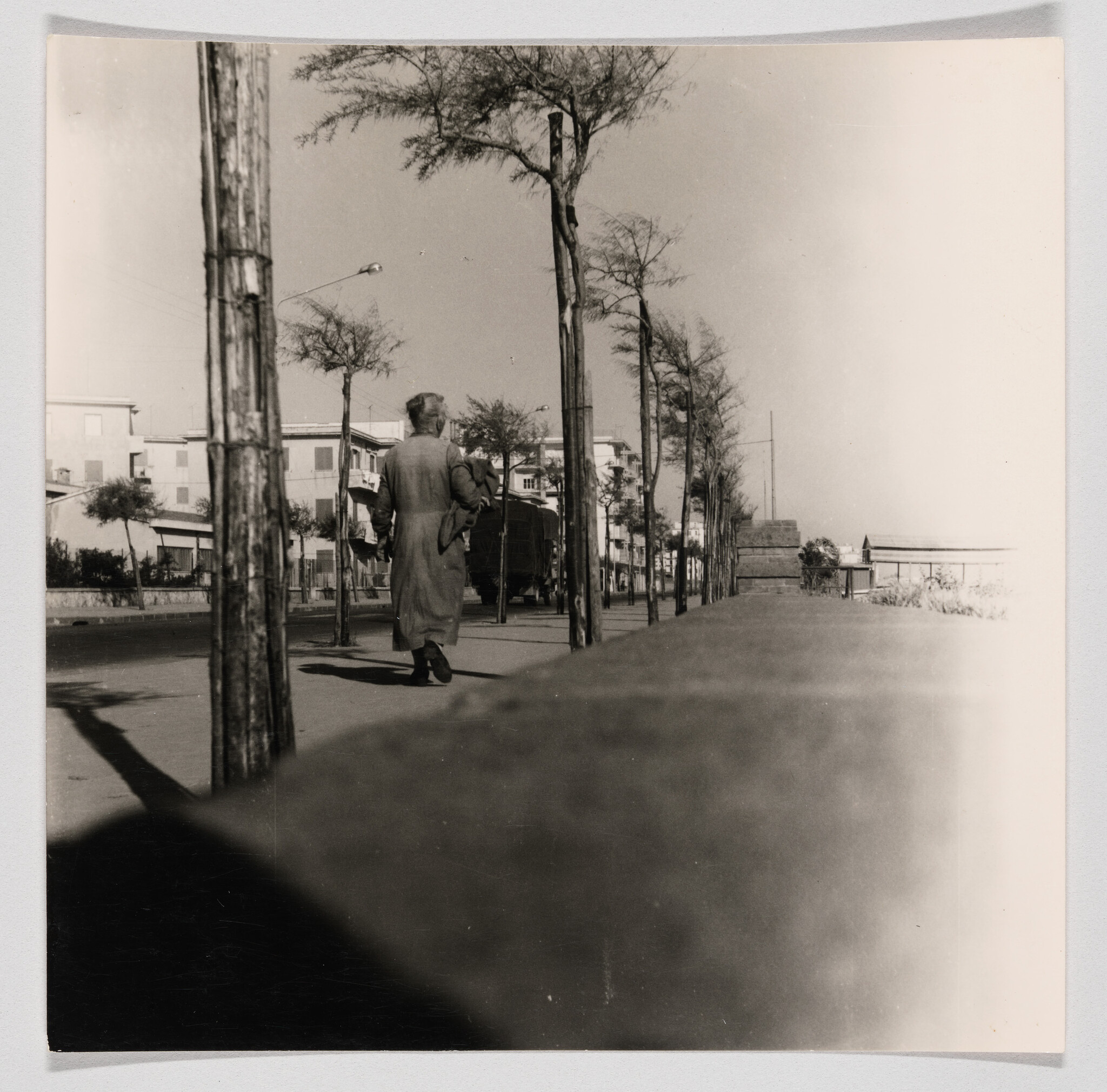 A person in a long coat walks down a tree-lined sidewalk away from the camera.