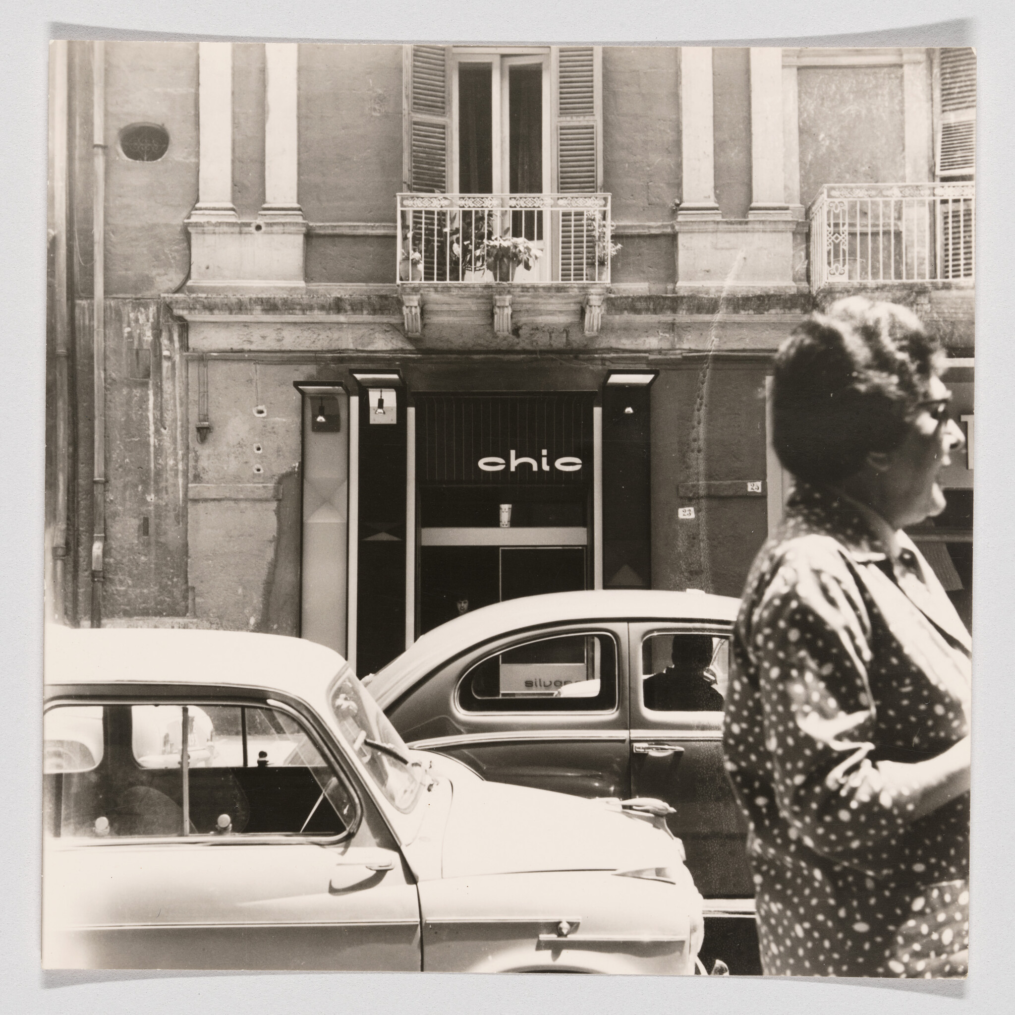 Woman walks past parked vintage cars in front of a building with a "chic" storefront.