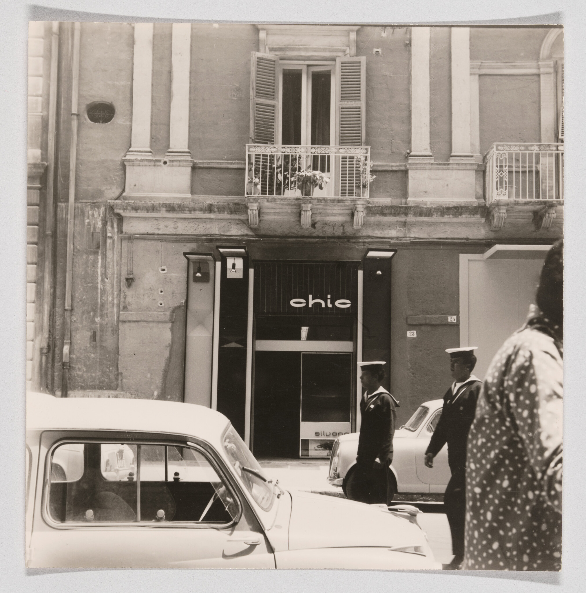 Two uniformed sailors walk past parked cars in front of a storefront labeled "chio".