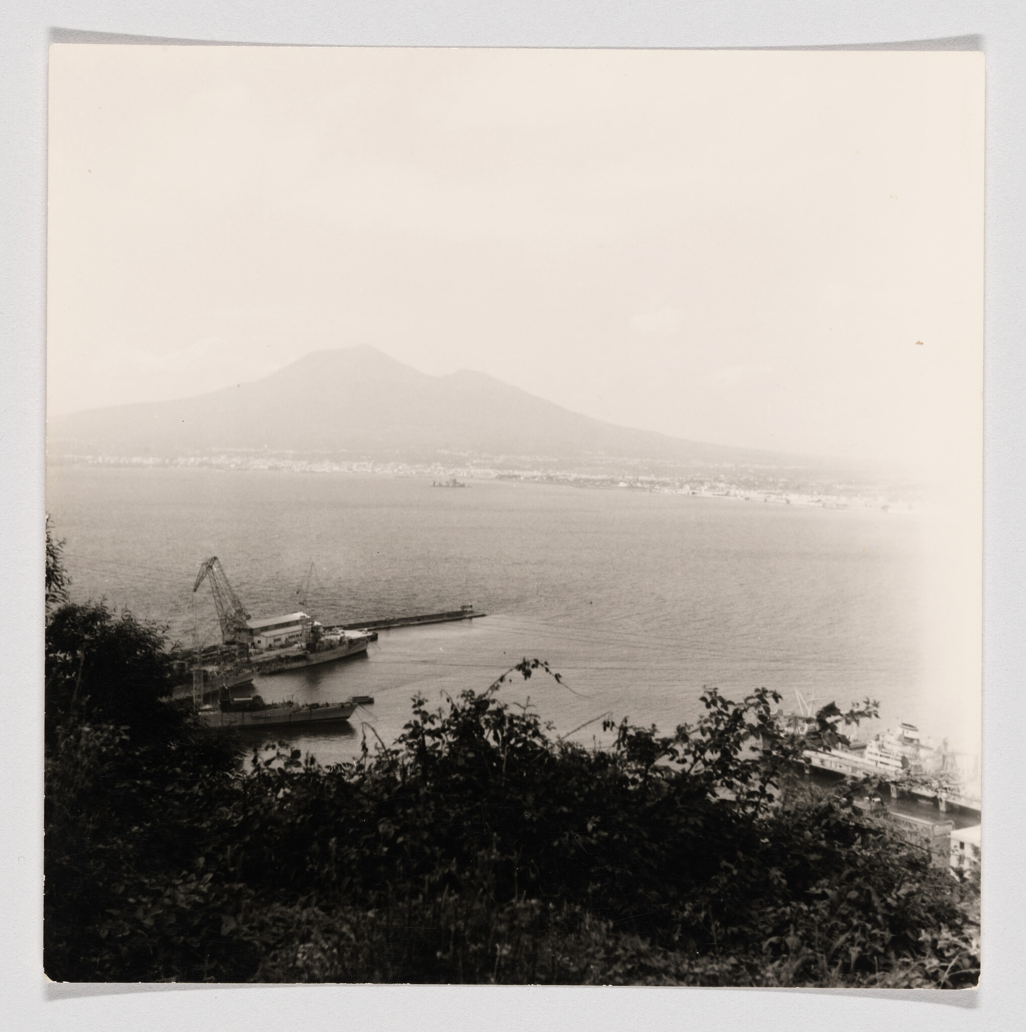 Harbor with cranes and ships in foreground and a large mountain rising across the calm bay.