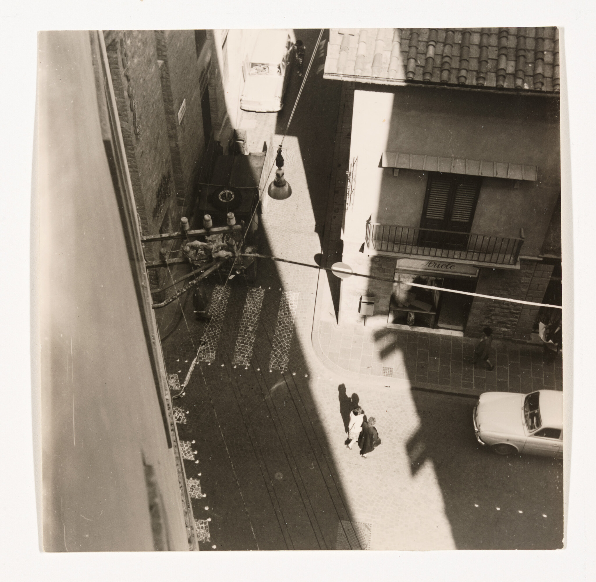 Two people walk together along a sunlit narrow street beside a parked car and building.