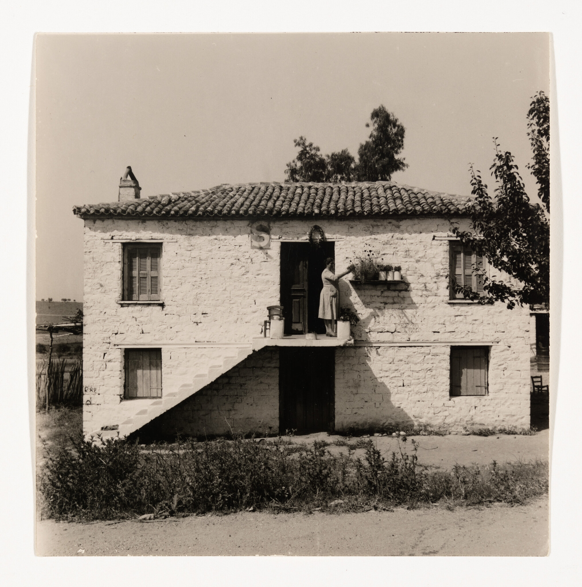 A woman waters potted plants on a second-floor ledge of a small stone house.