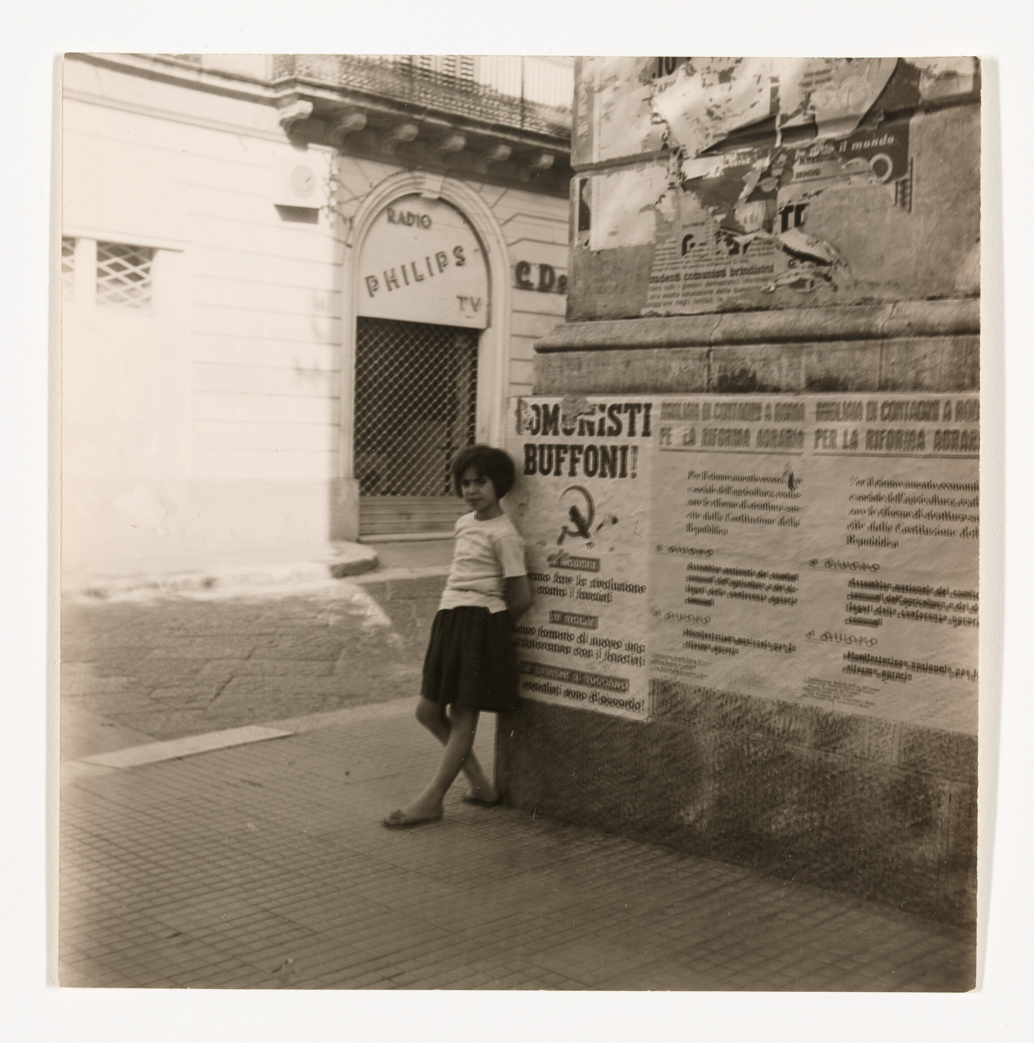 A young girl leans against a corner wall covered in political posters near a Philips store.