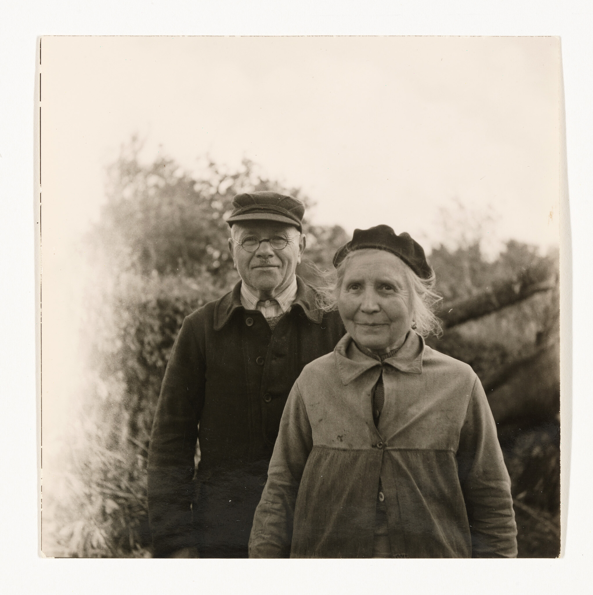 An older man and woman stand together outdoors, both wearing coats and caps, looking at the camera.
