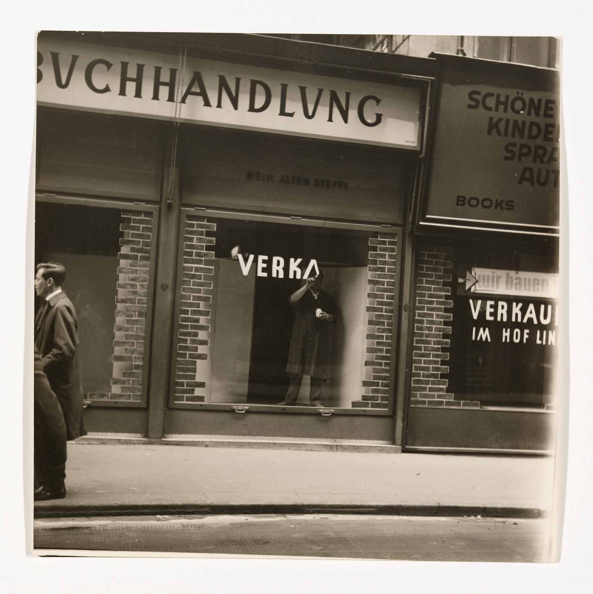 Man stands inside a bookstore window adjusting his hat while a passerby walks by outside.