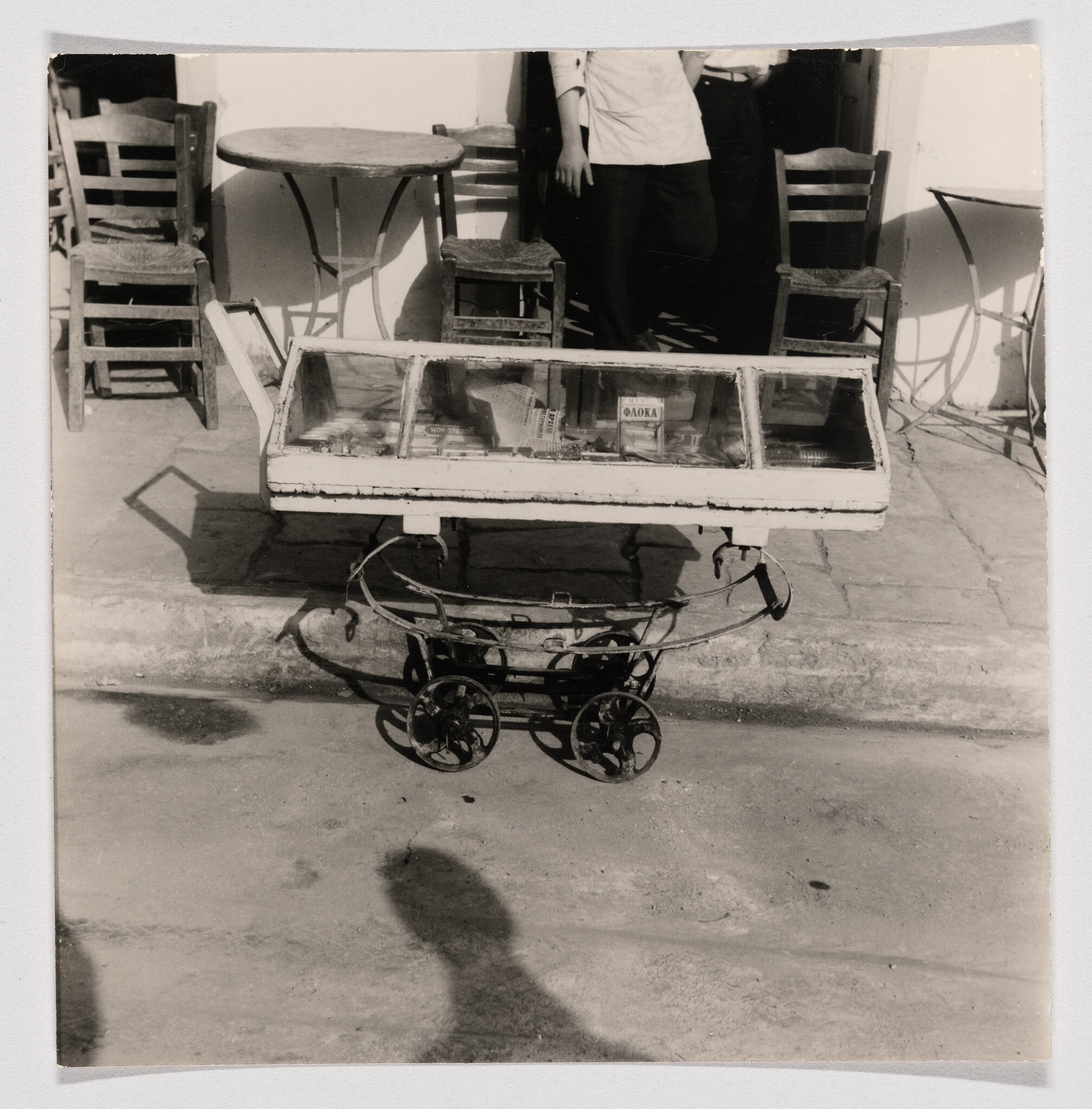 A small wheeled glass display cart filled with goods parked in front of a cafe with chairs.