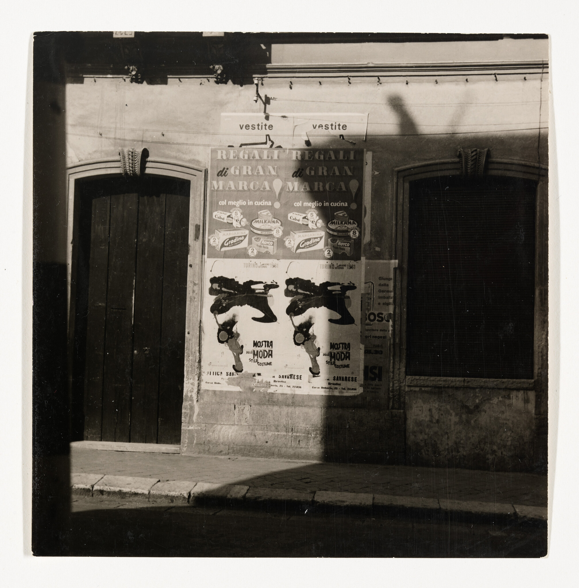 Weathered wall between two arched doors displays layered posters casting a tall shadow on the sidewalk.