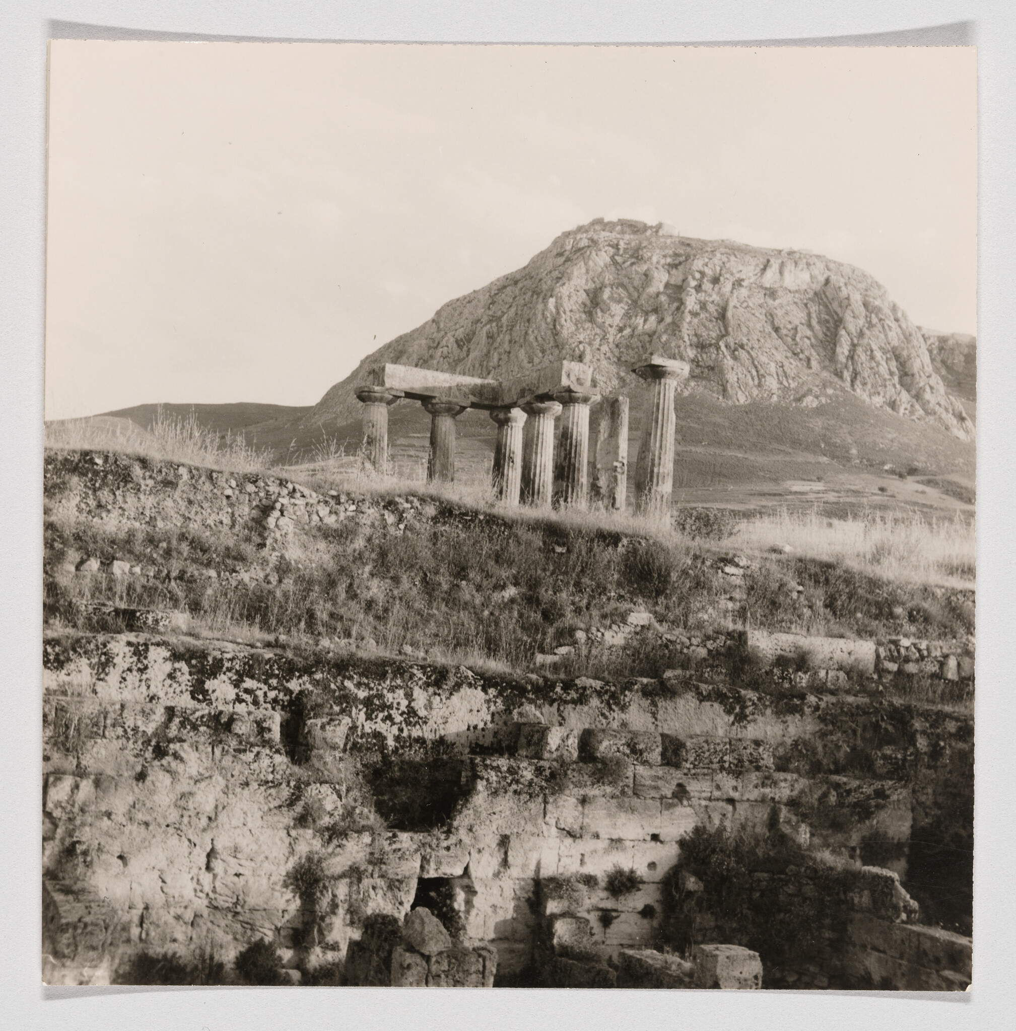 Ruined Greek temple columns stand on a grassy hillside with a large rocky mountain behind.
