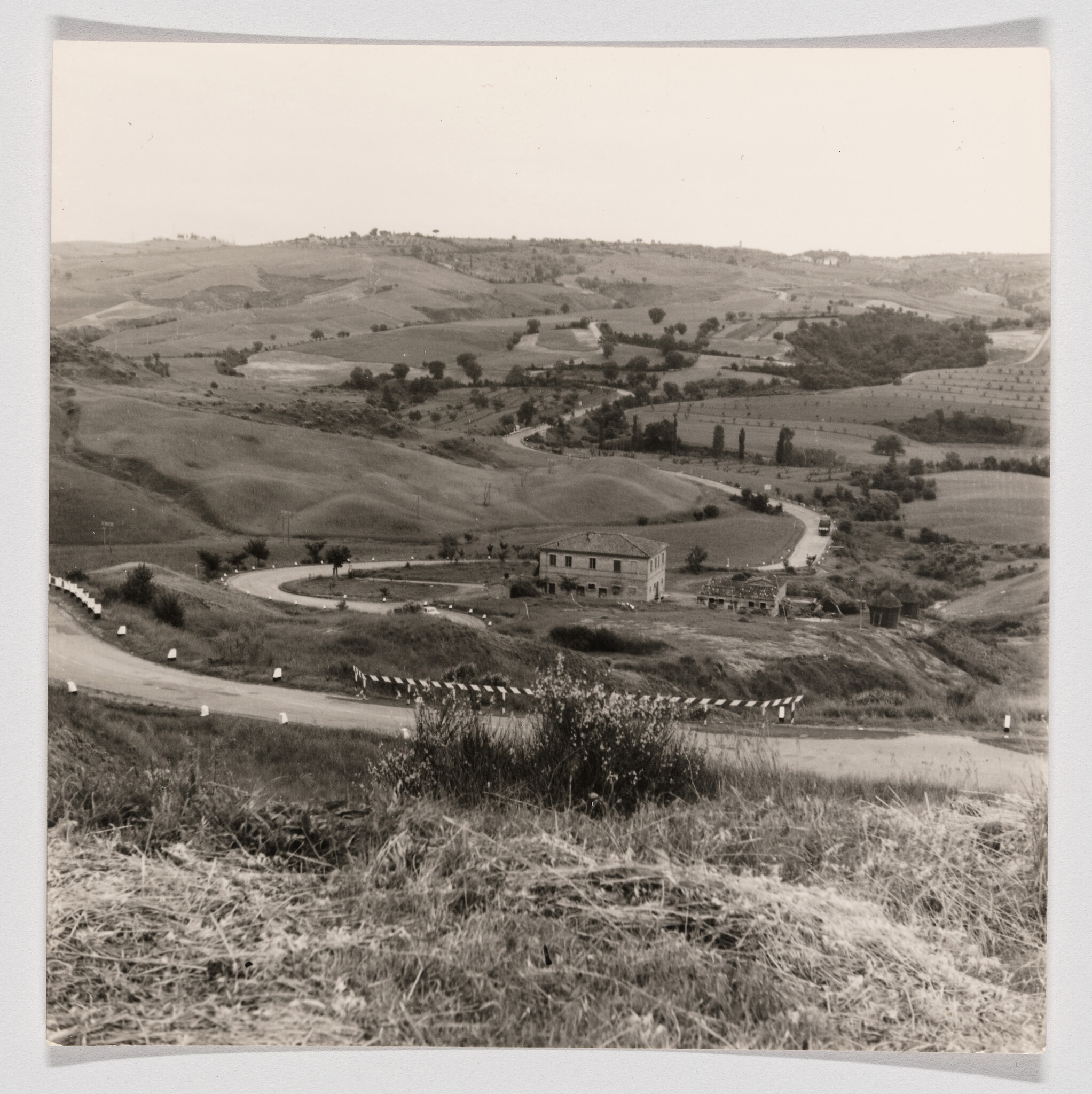 A winding rural road curves through rolling hills past a solitary farmhouse and fields.