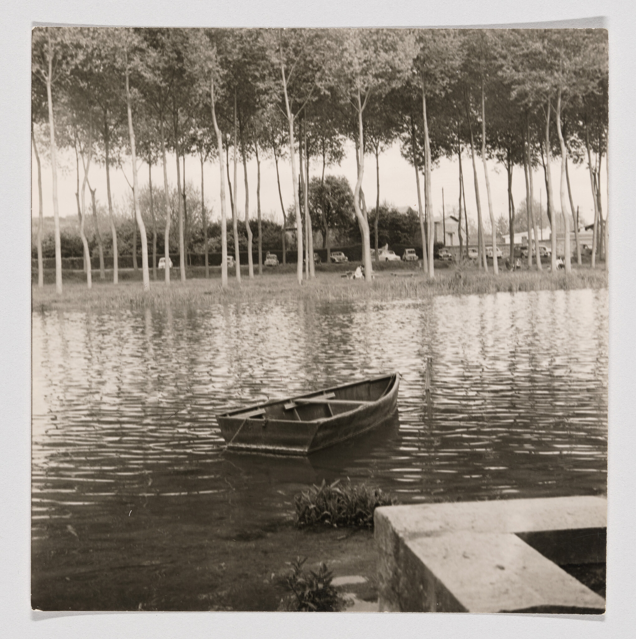 A small wooden rowboat tied near the shore of a calm river with trees and parked cars beyond.