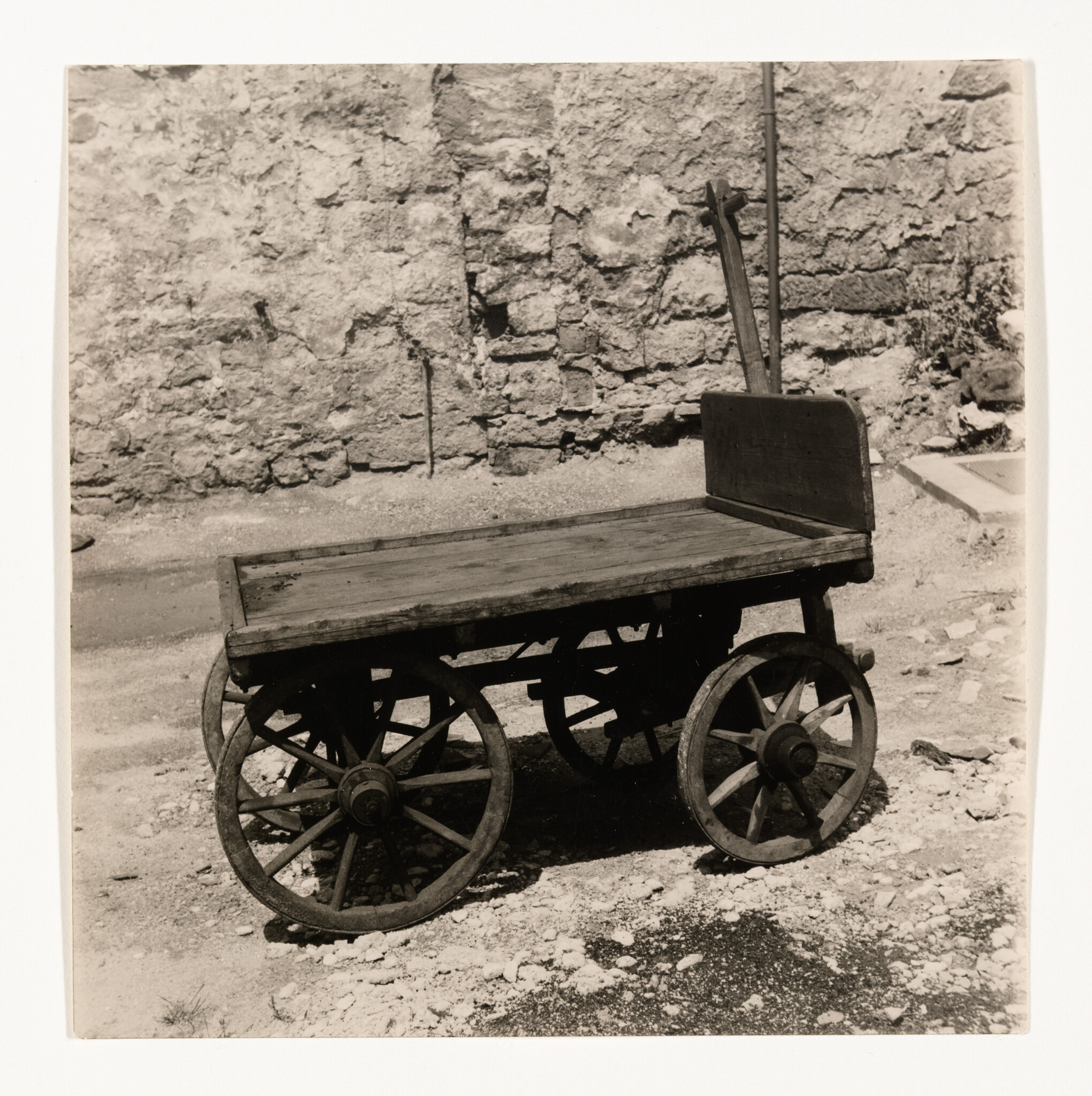 Old wooden flatbed cart with four spoked wheels sits on rough ground near a stone wall.