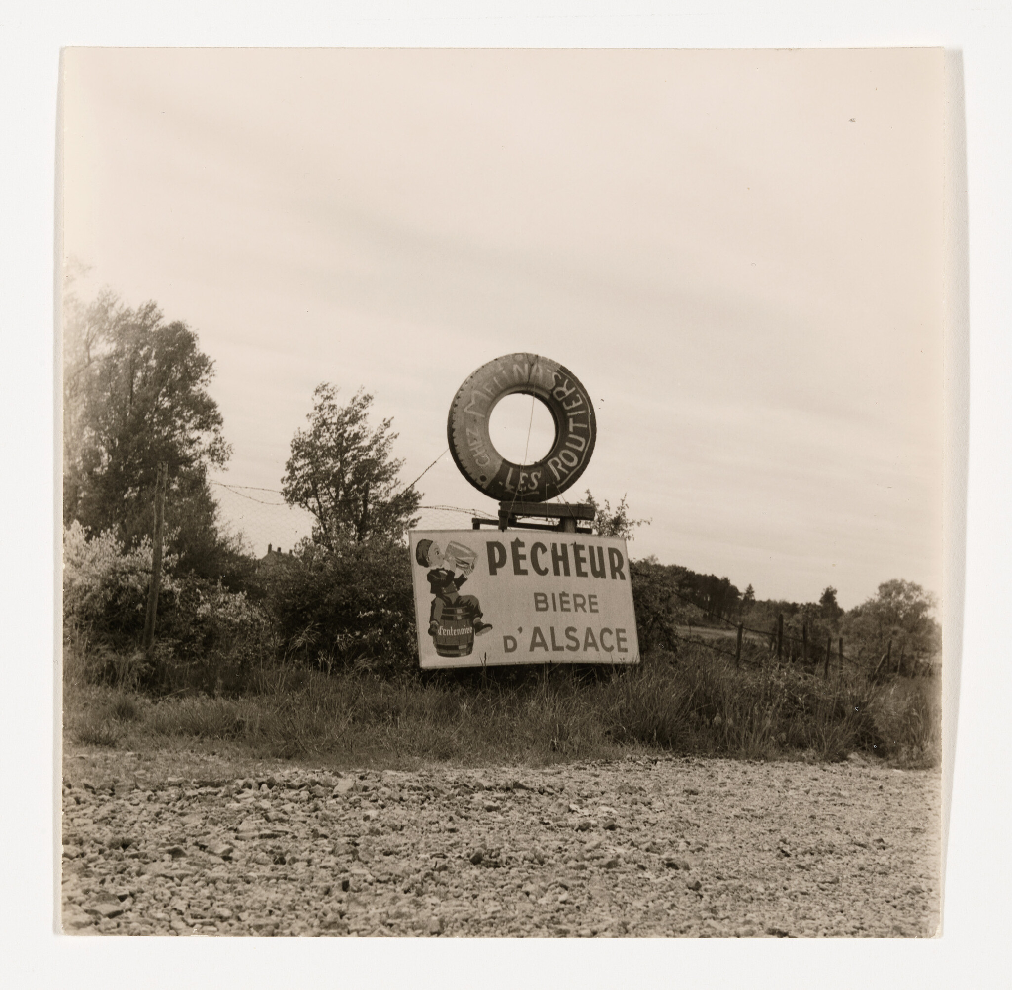 A vintage black and white photograph depicting an old roadside advertisement sign for "Pêcheur Biere d'Alsace" with a large graphic of a fisherman and a tire mounted above the sign. The setting appears rural with trees and a fence in the background, and the ground in the foreground is rocky and uneven.
