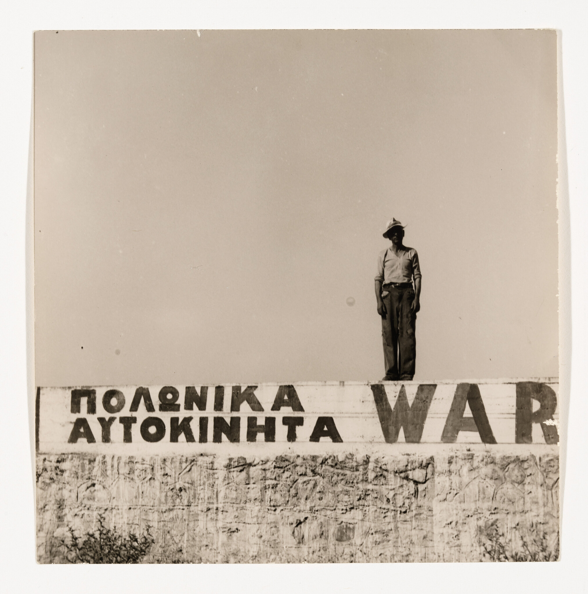 Man wearing a hat stands on top of a painted wall with Greek text and the word WAR.