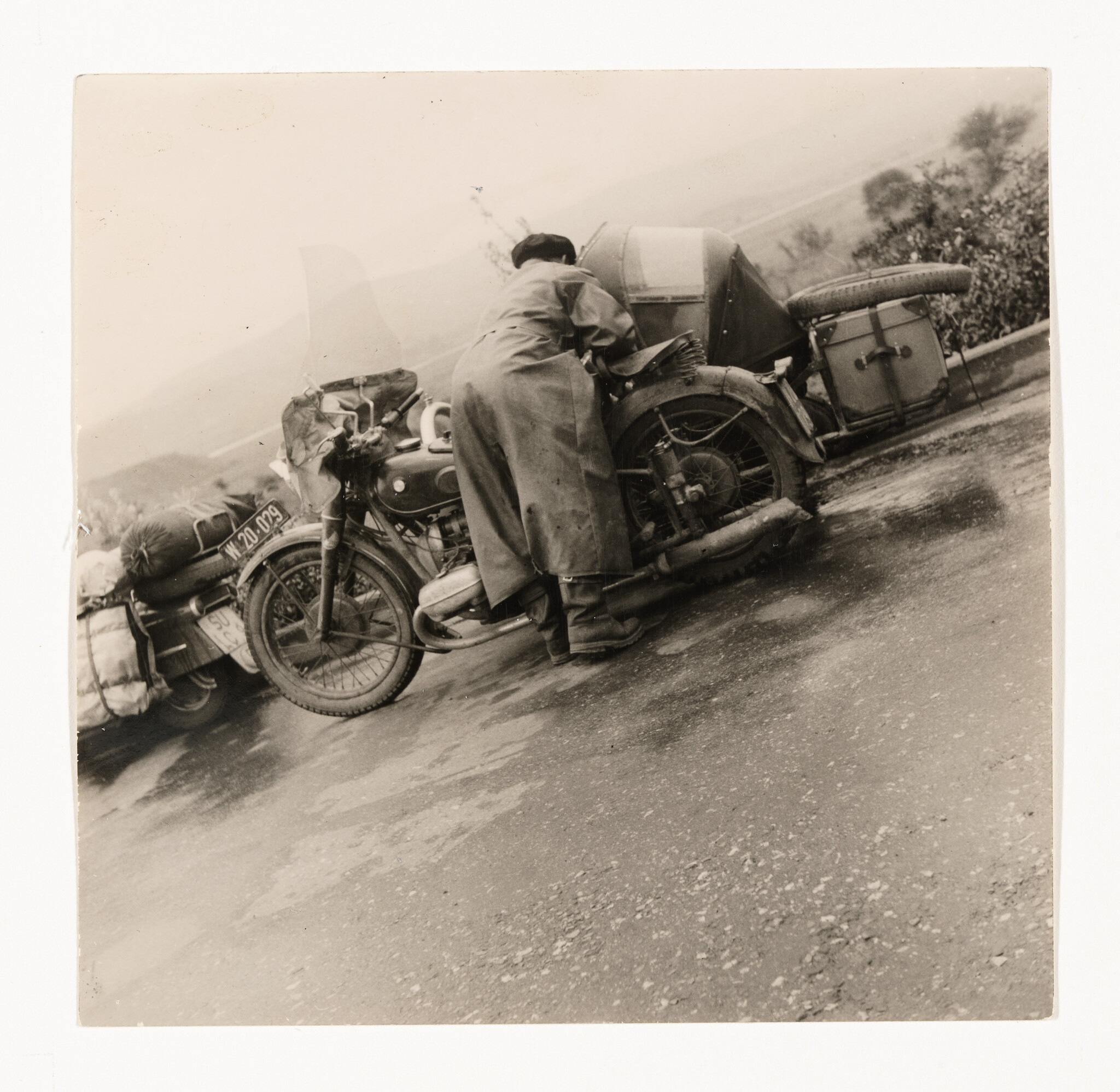 A person in a long coat leans over a motorcycle with sidecar on a wet road.