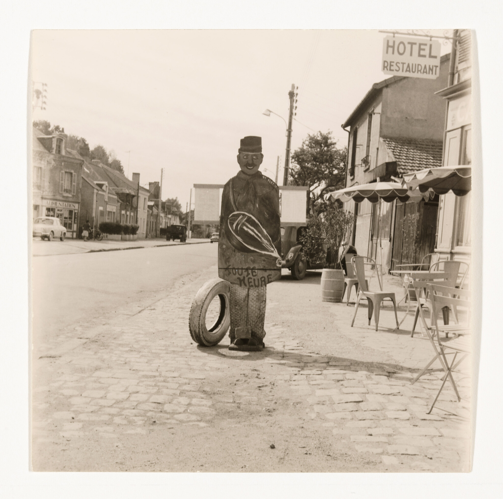 A painted wooden figure stands on a cobblestone street next to a tire outside a hotel restaurant.
