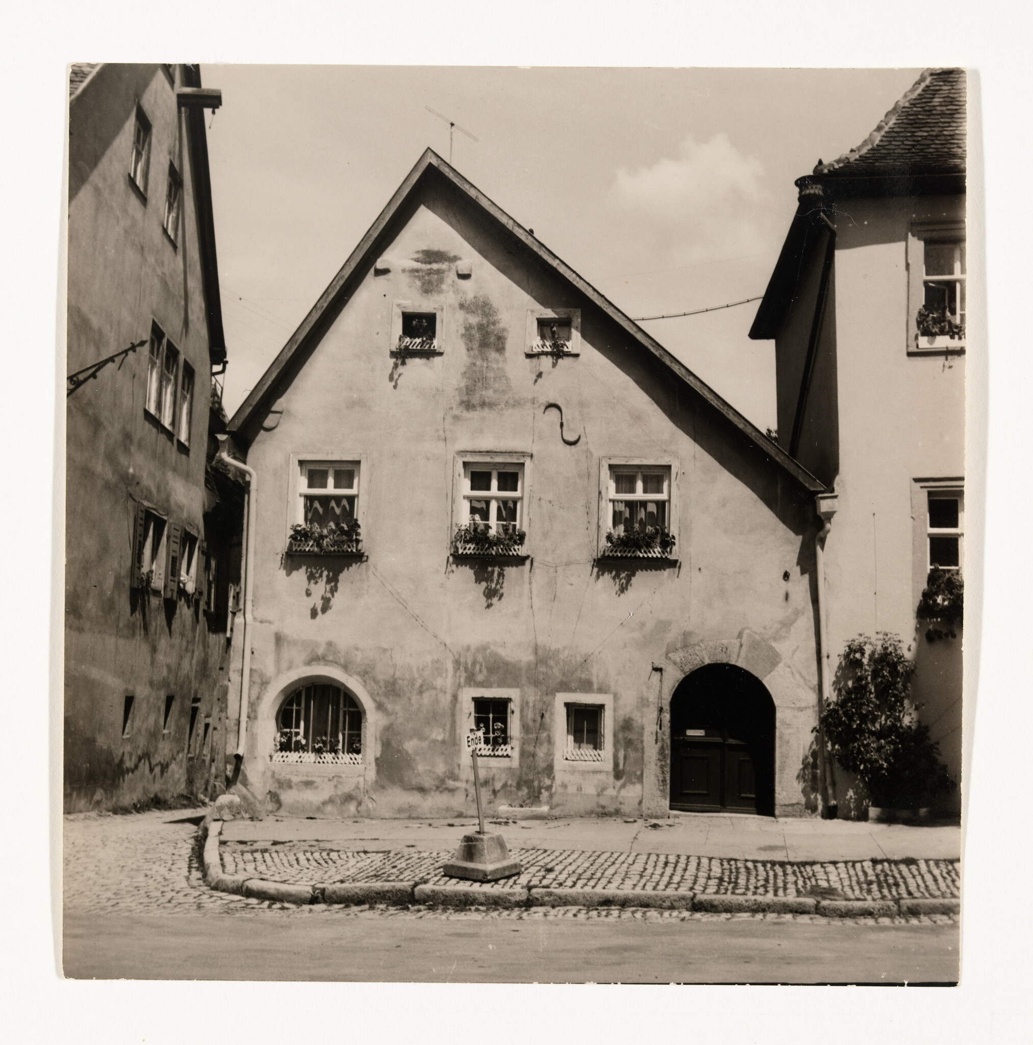 A small old house with flower-filled window boxes, an arched doorway, and a cobblestone street.