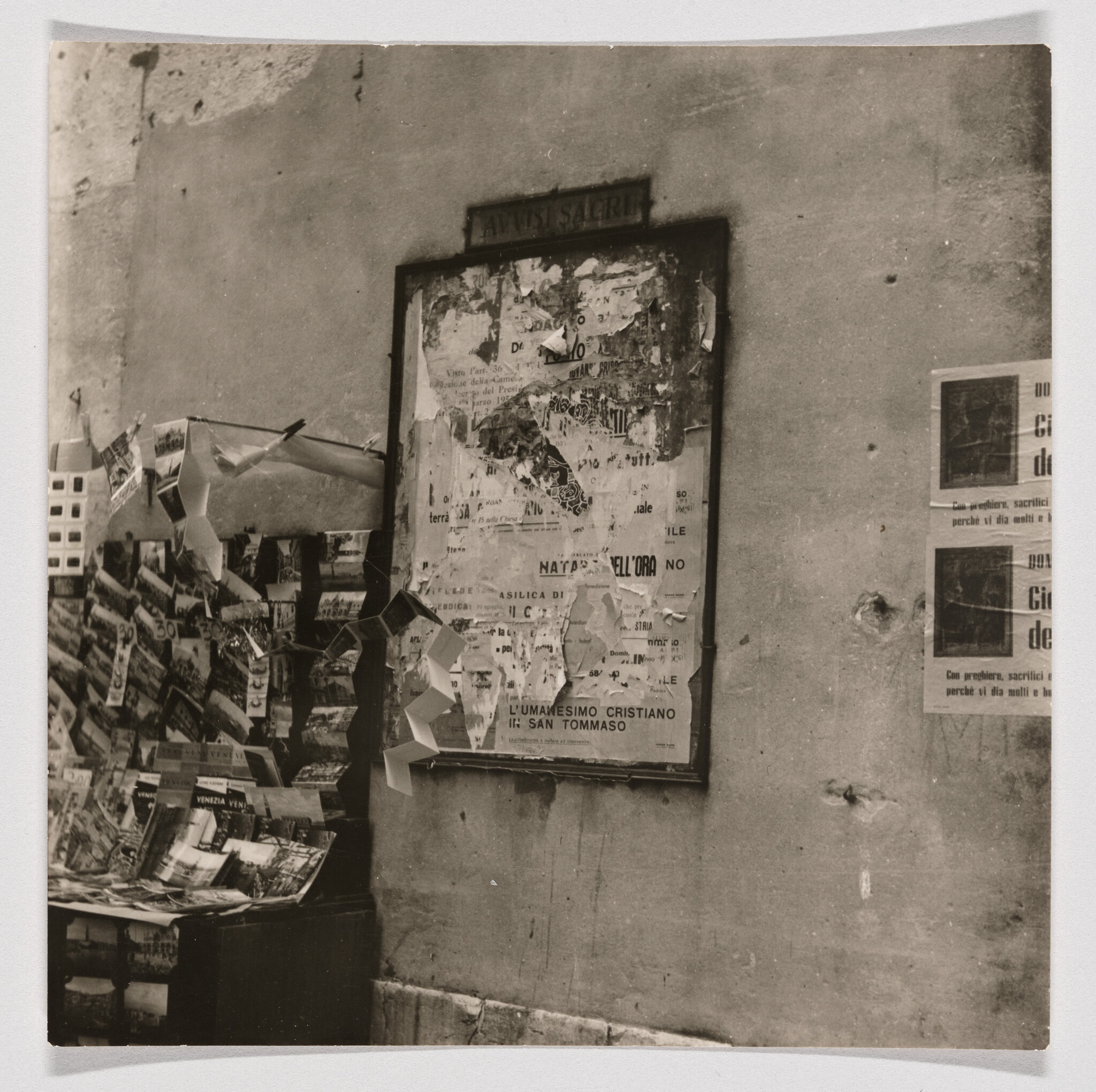 Torn poster board on a wall next to a street newsstand displaying postcards and pamphlets.