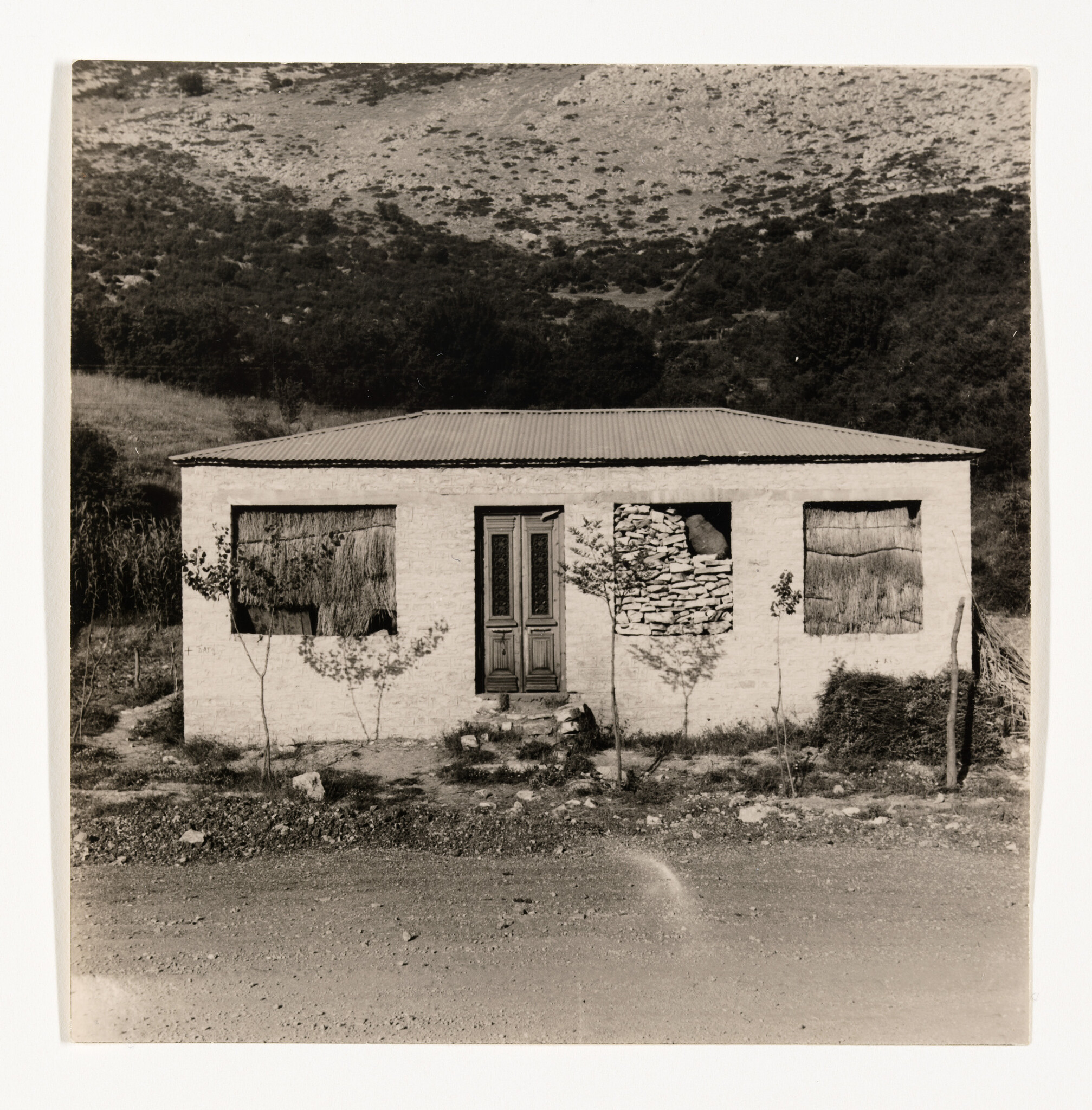 Small abandoned brick house with boarded windows and central double door in rural landscape.