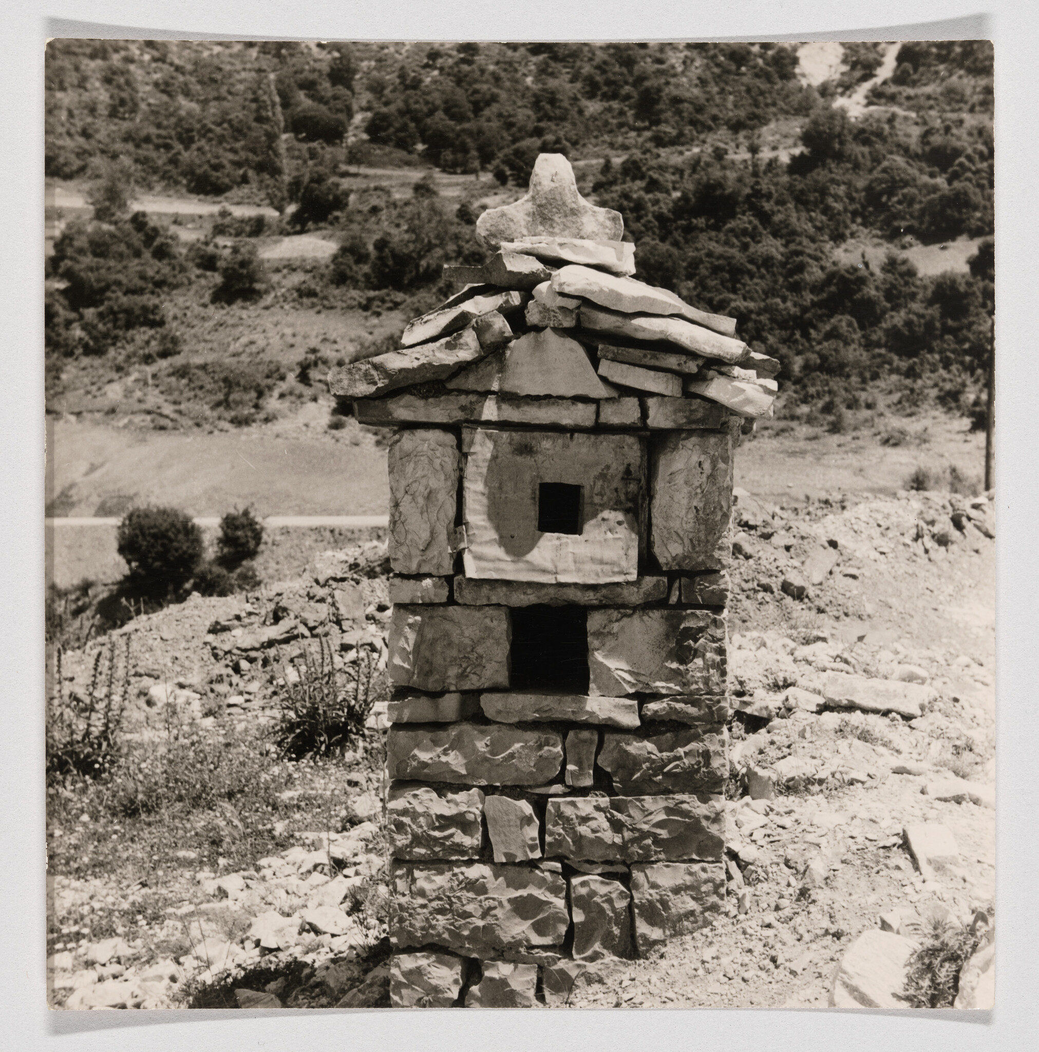Small stacked stone shrine with two square openings stands on a rocky hillside under trees.