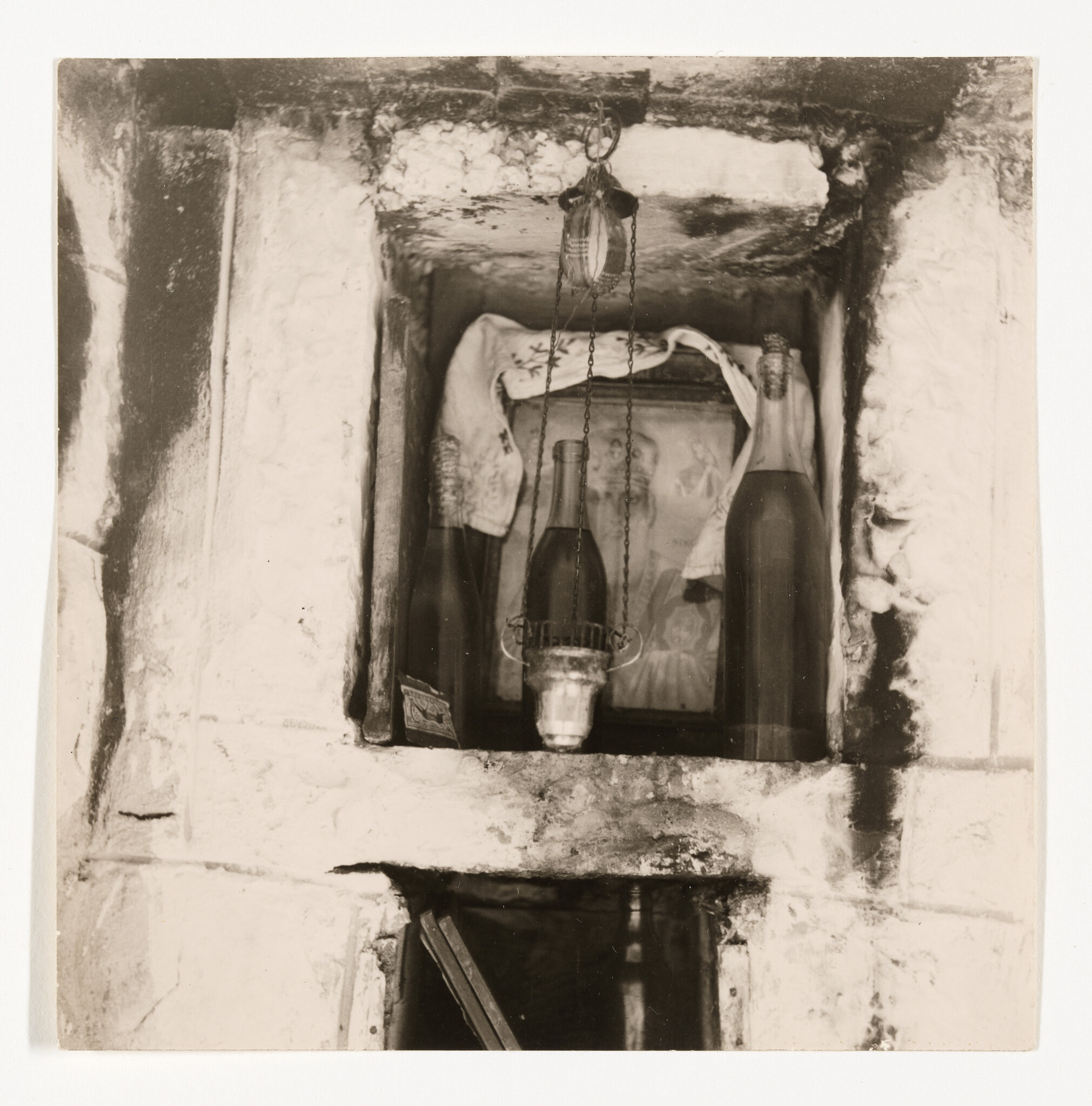 A small wall shrine niche holding two bottles, a hanging oil lamp, and a draped cloth.