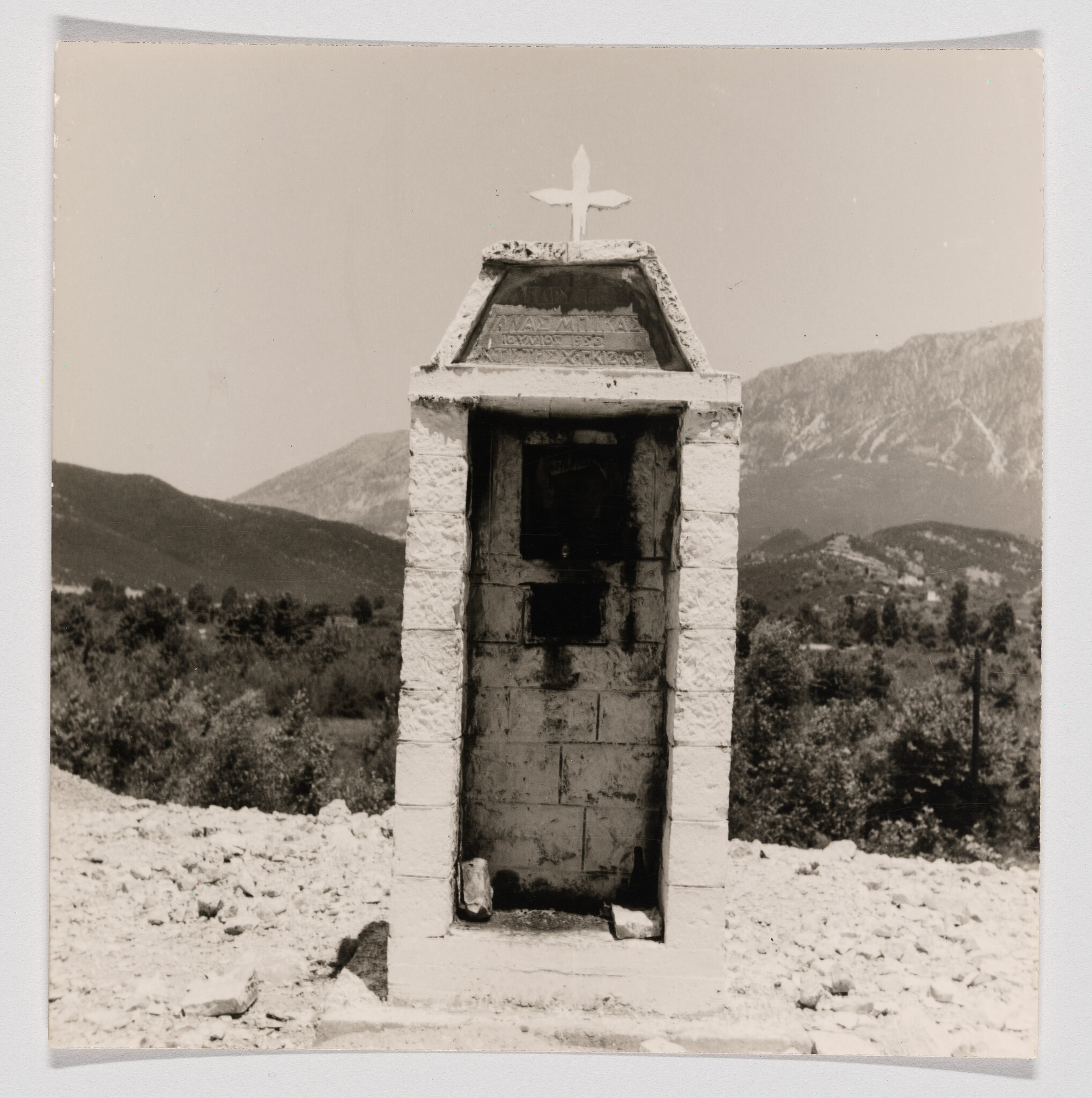 A small stone roadside shrine with a cross stands empty against a mountainous landscape.