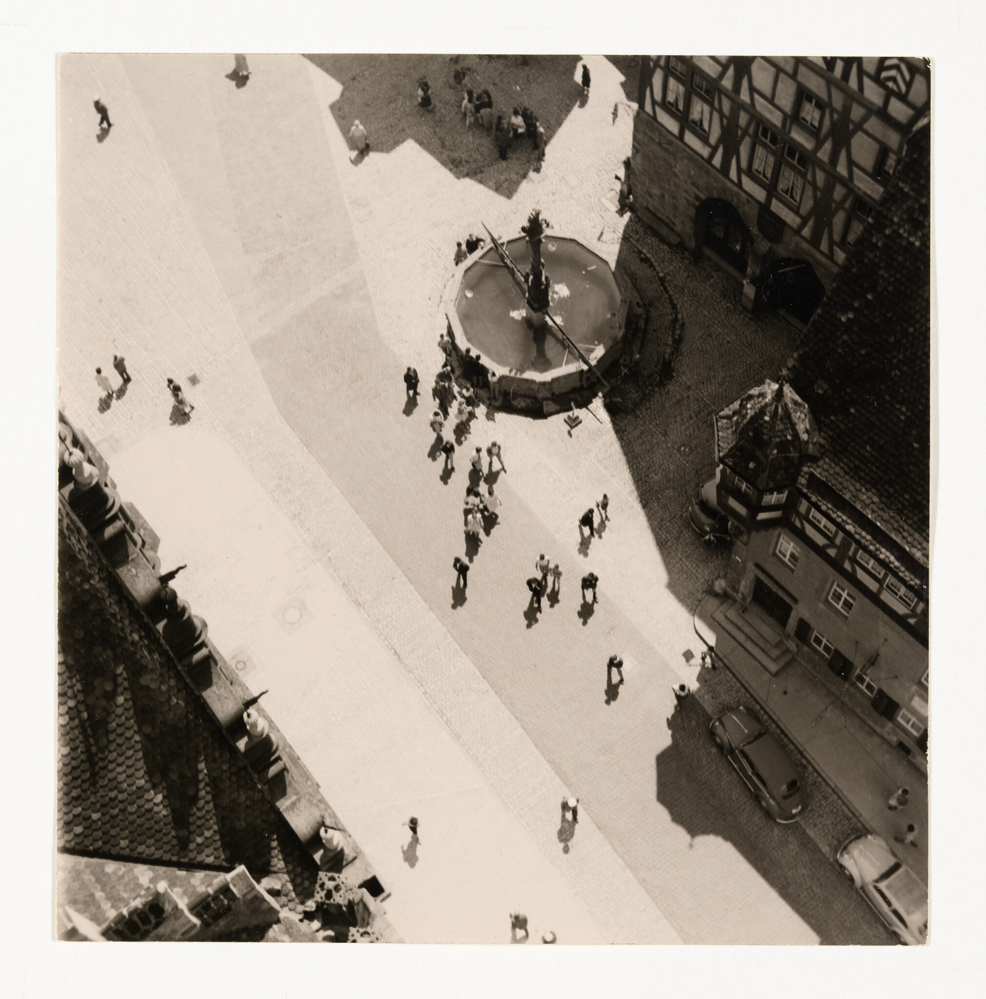 People walk around a central fountain in a sunlit town square seen from above.