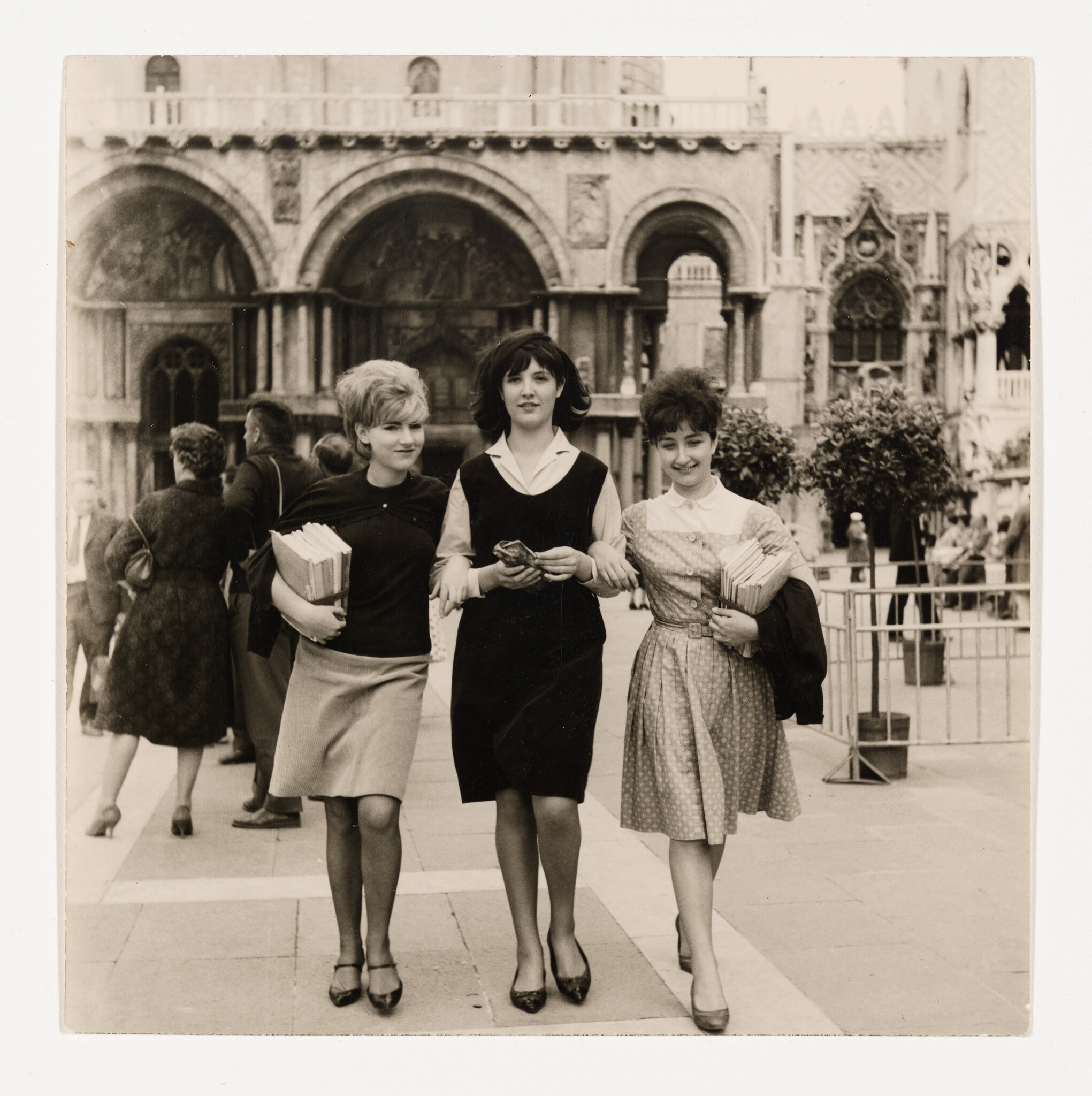 Three young women walk arm in arm down a plaza holding books in front of an ornate building.