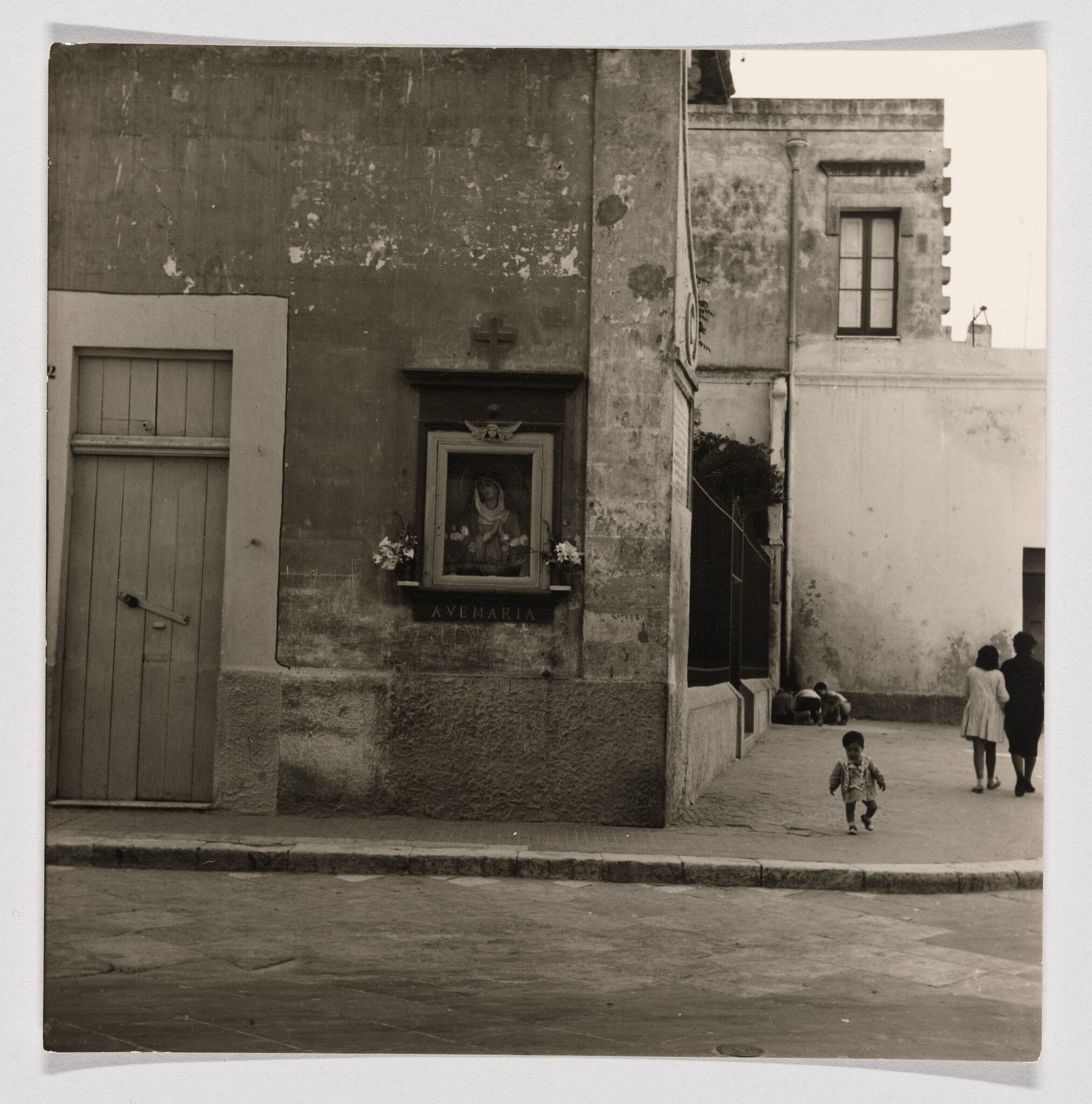 A small child walks past a wall shrine labeled "Ave Maria" on a quiet street corner.