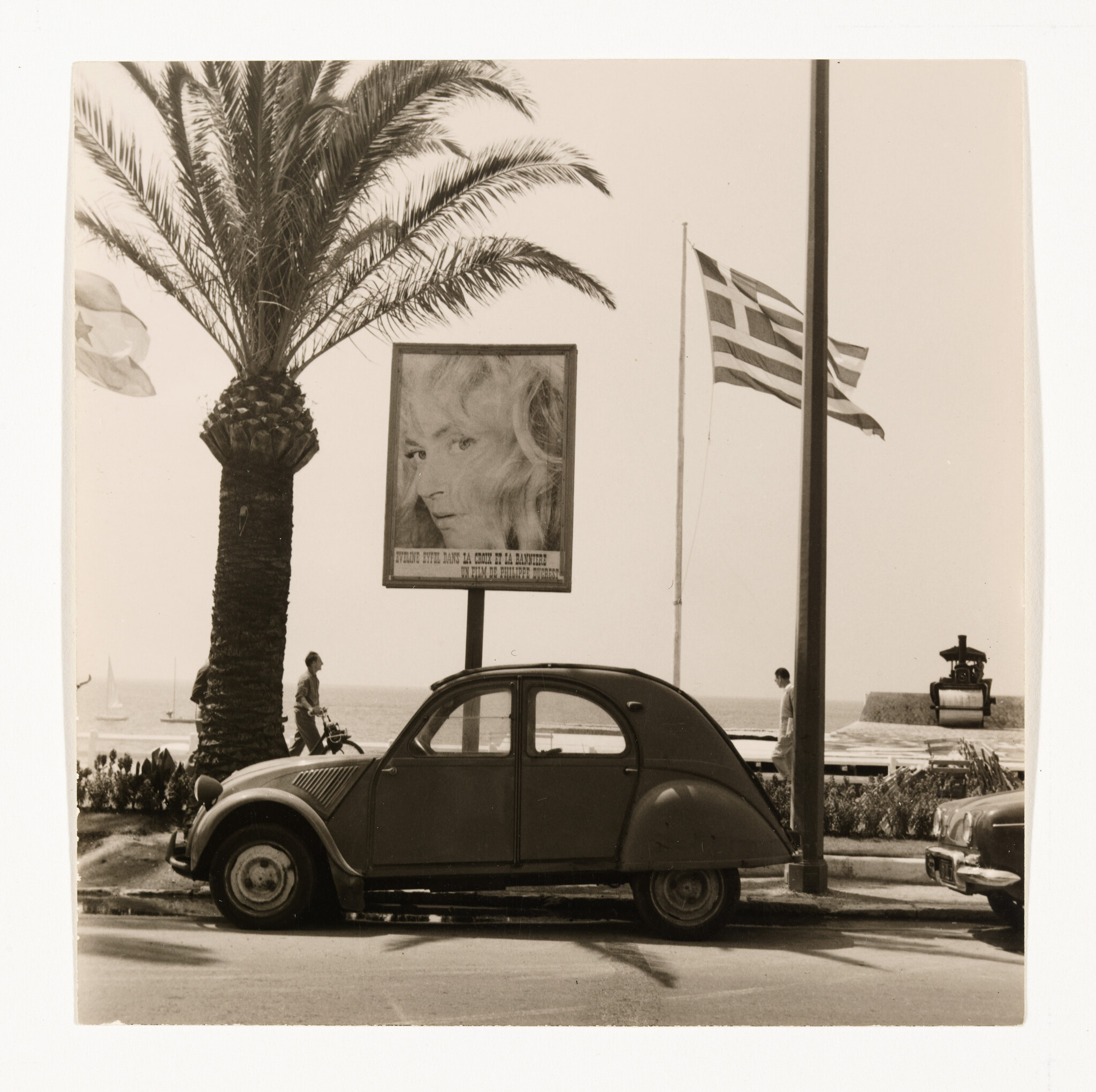 A vintage Citroën is parked by the seaside beneath a large billboard and Greek flag.