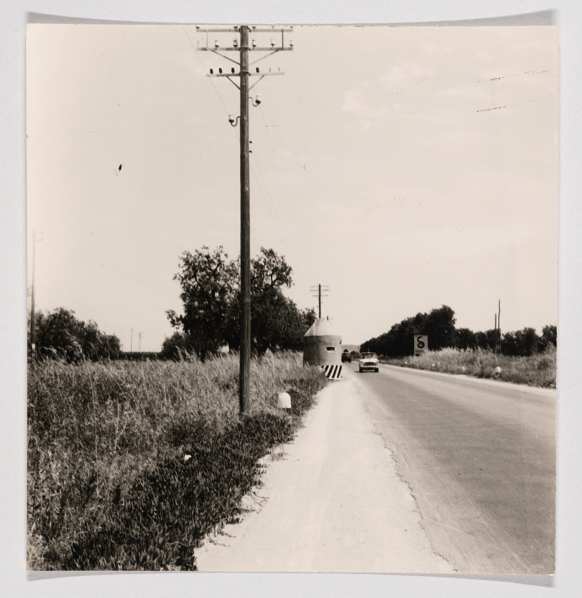 A car drives past a concrete roadside guard post near a telephone pole on a country road.