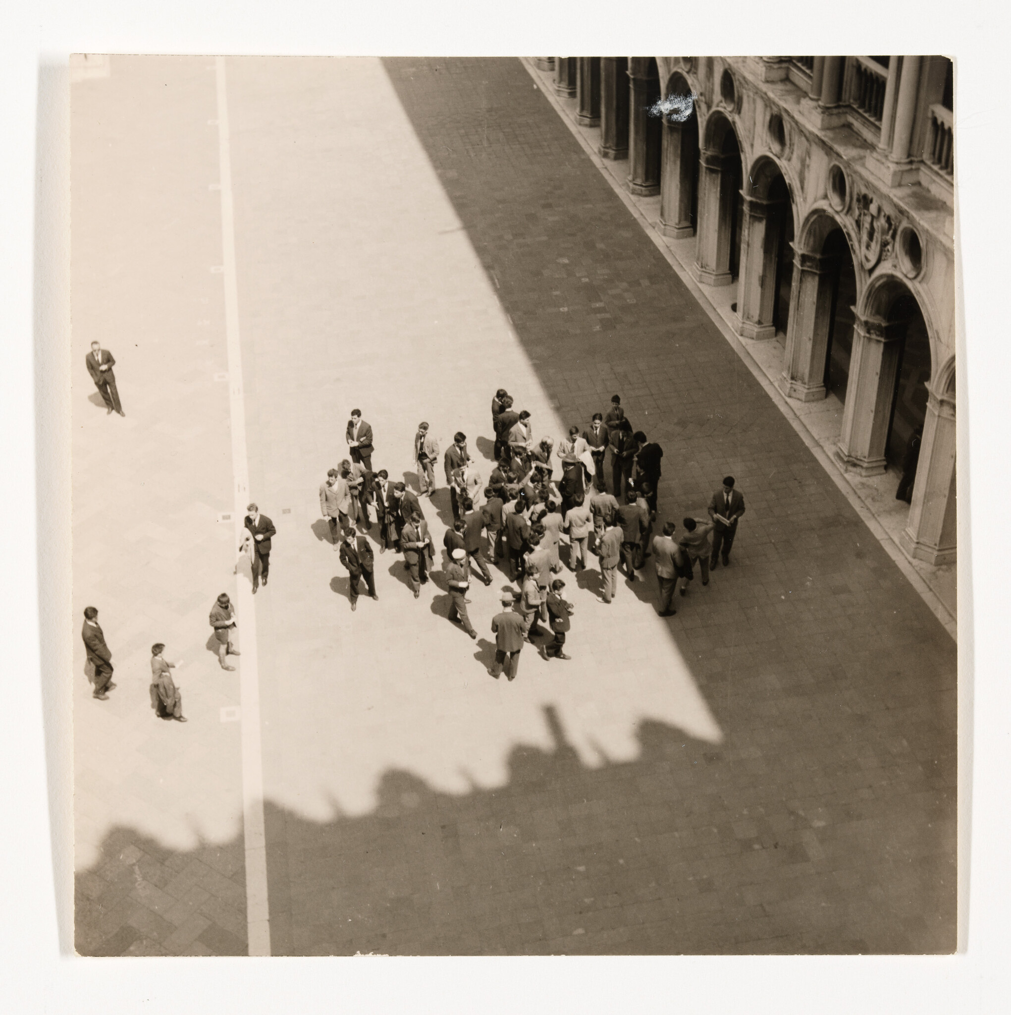 A group of people gathers in a sunlit plaza beside an arched building, with a long shadow across the square.