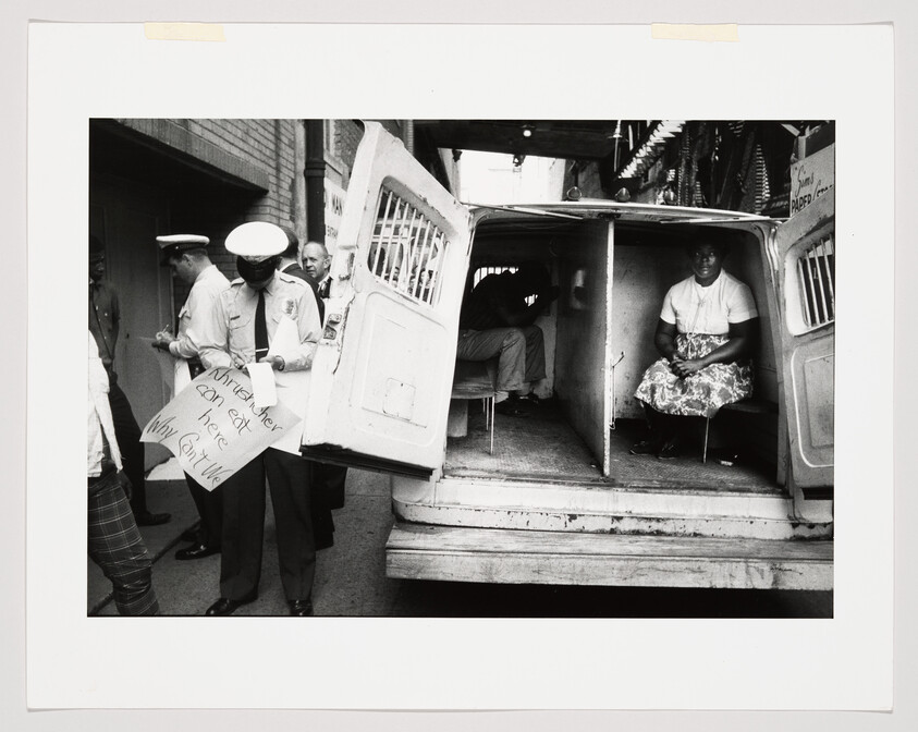Police officer writes a citation while a woman sits inside a divided police van with doors open.