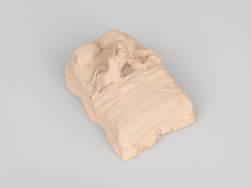 A small beige clay sculpture of a person sitting up in bed with a wrinkled blanket.