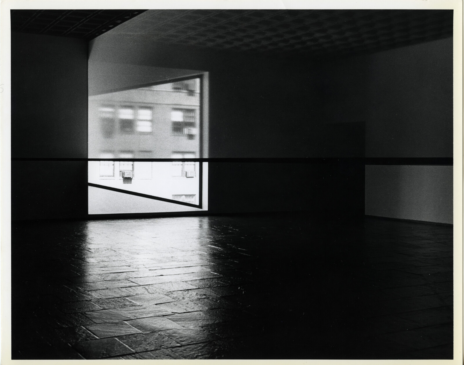 Empty gallery room with a bright window, diagonal handrail, and light reflected on the tiled floor.