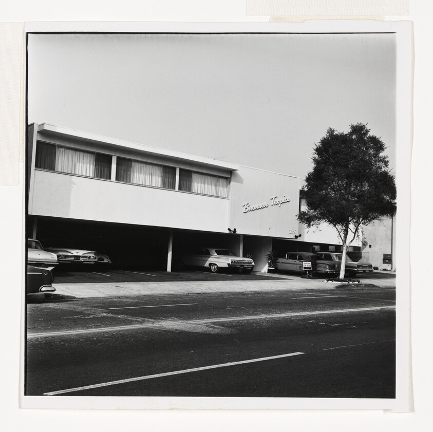 Two-story motel with covered parking and several vintage cars along a quiet street.