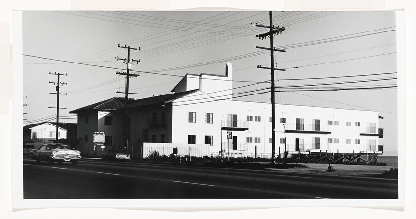 Large white apartment building beside a busy road with utility poles and passing cars.