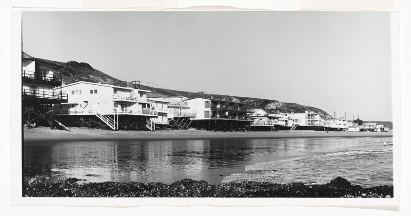 A row of stilted beachfront houses lines the calm shoreline under a clear sky.