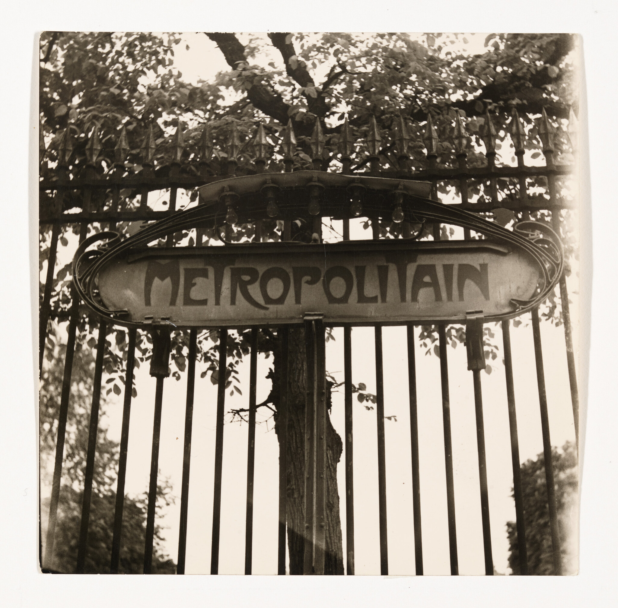 An ornate Metropolitain sign hangs above a tall iron gate with tree branches behind it.