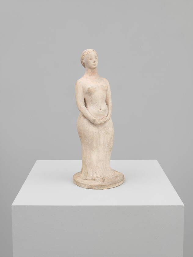 Small pale clay sculpture of a standing woman with hands clasped on a white pedestal.