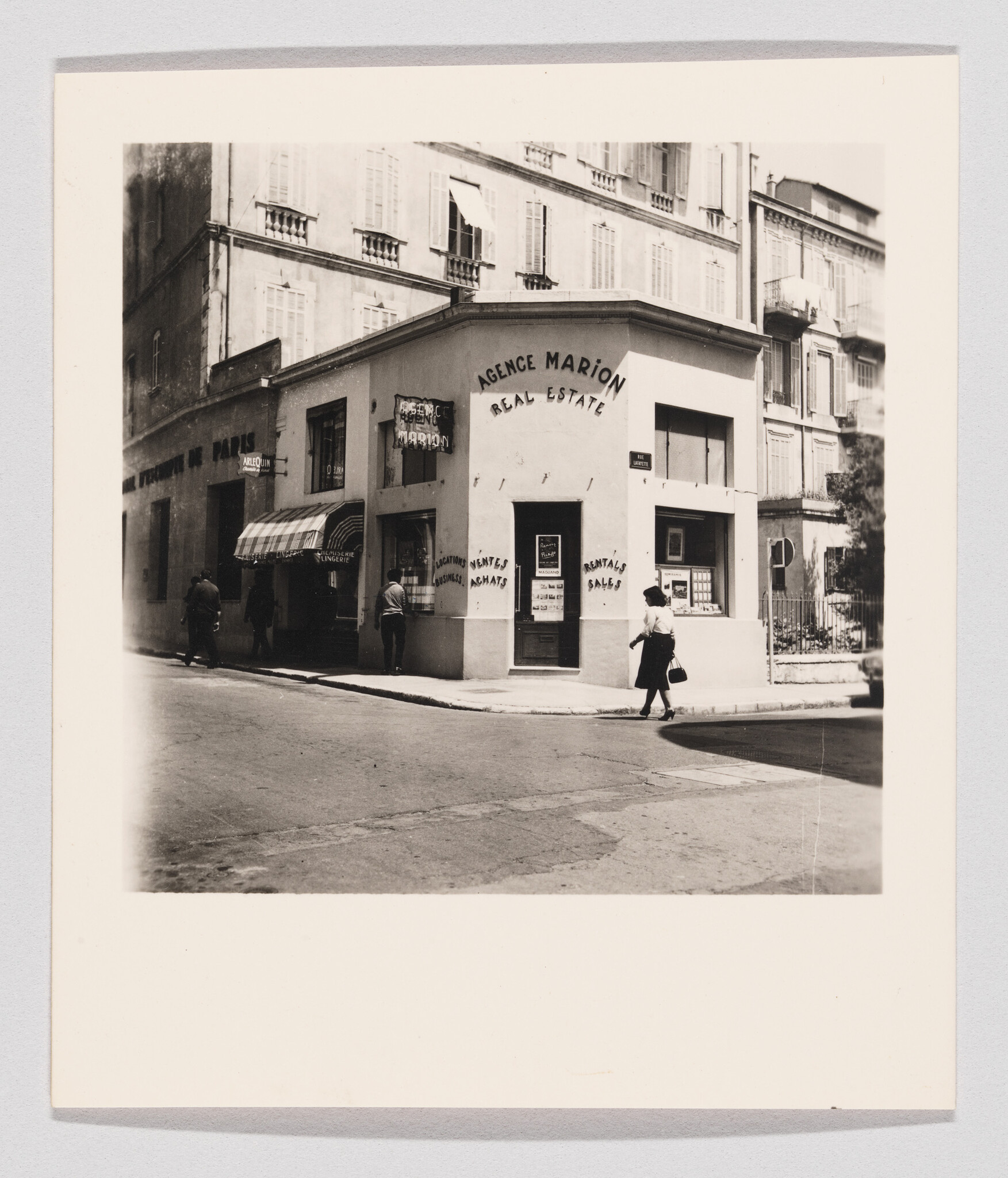 A woman walks past a corner real estate office labeled 'Agence Marion' in a city.