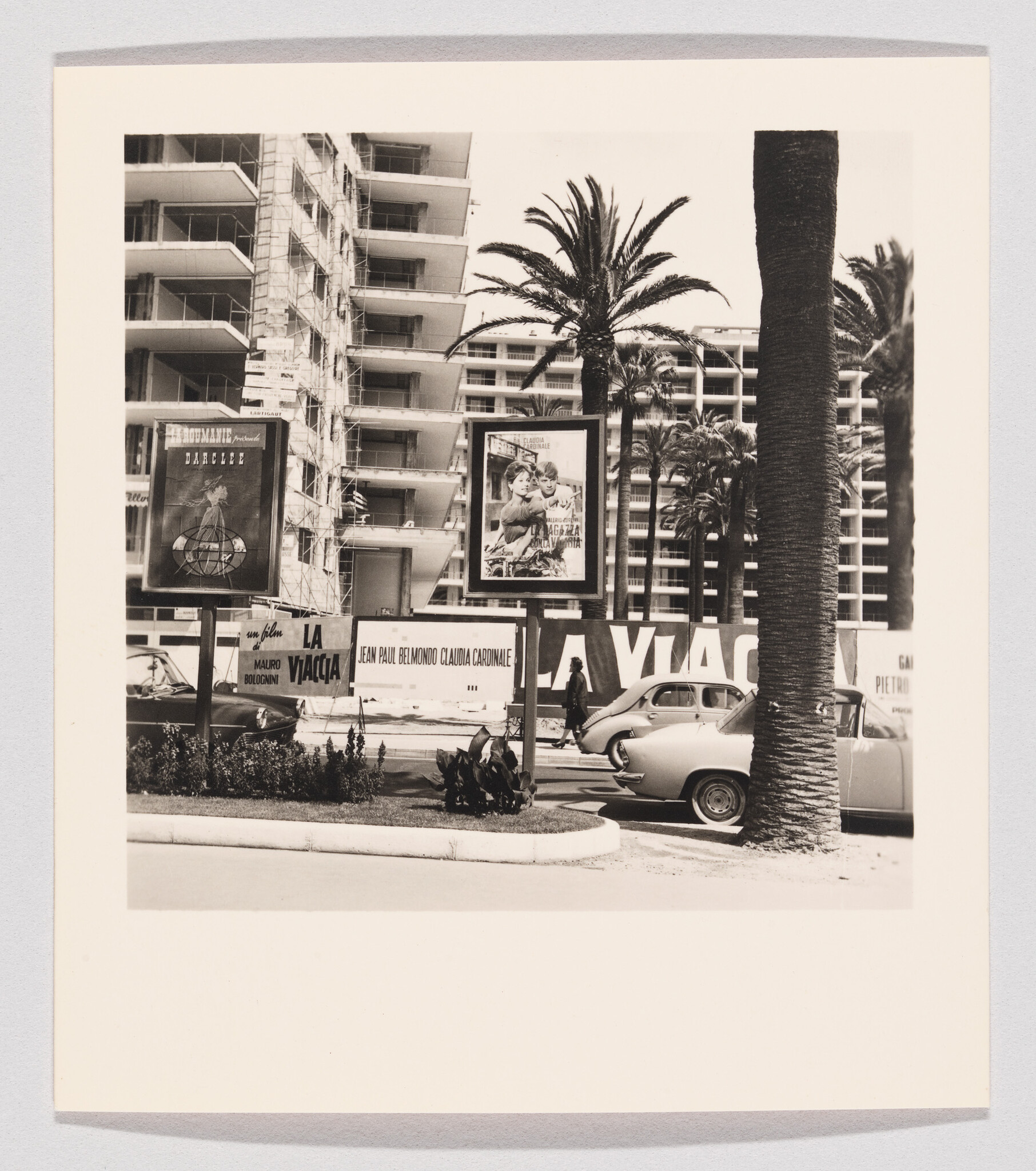 Palm-lined street with vintage cars parked beside movie posters and a modernist apartment building.
