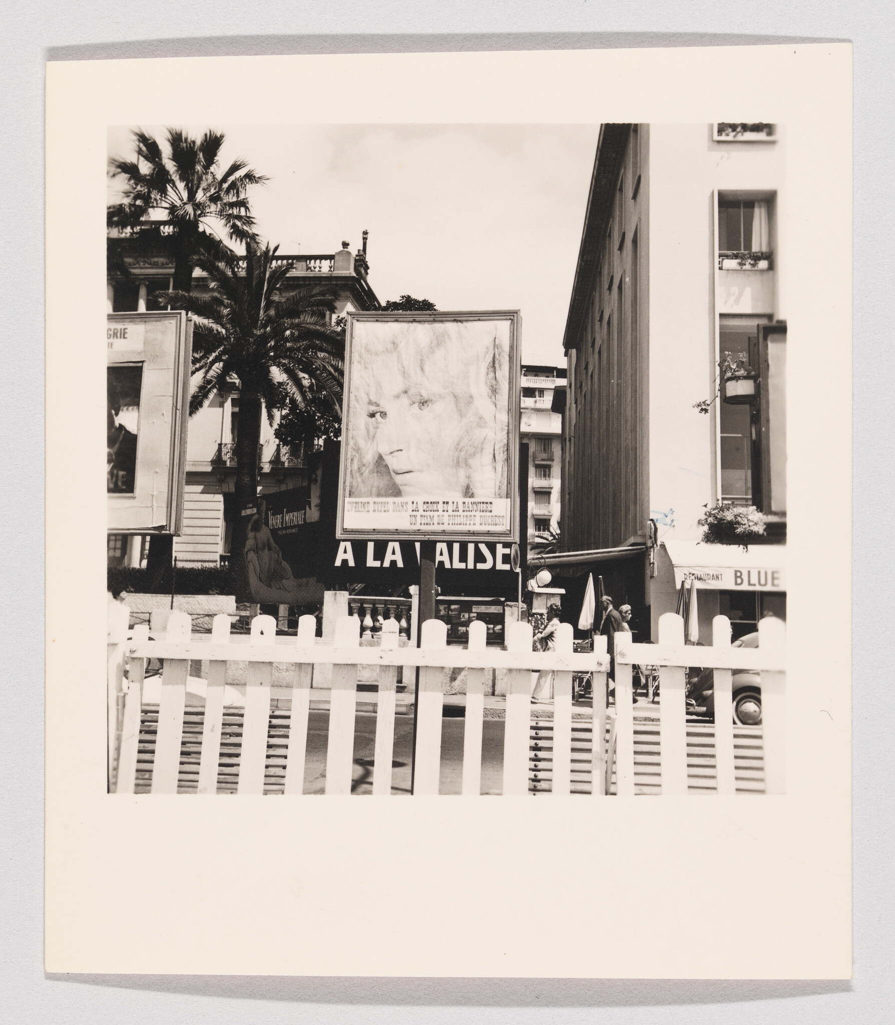 Large billboard with a woman's face stands behind a white picket fence in a city street.