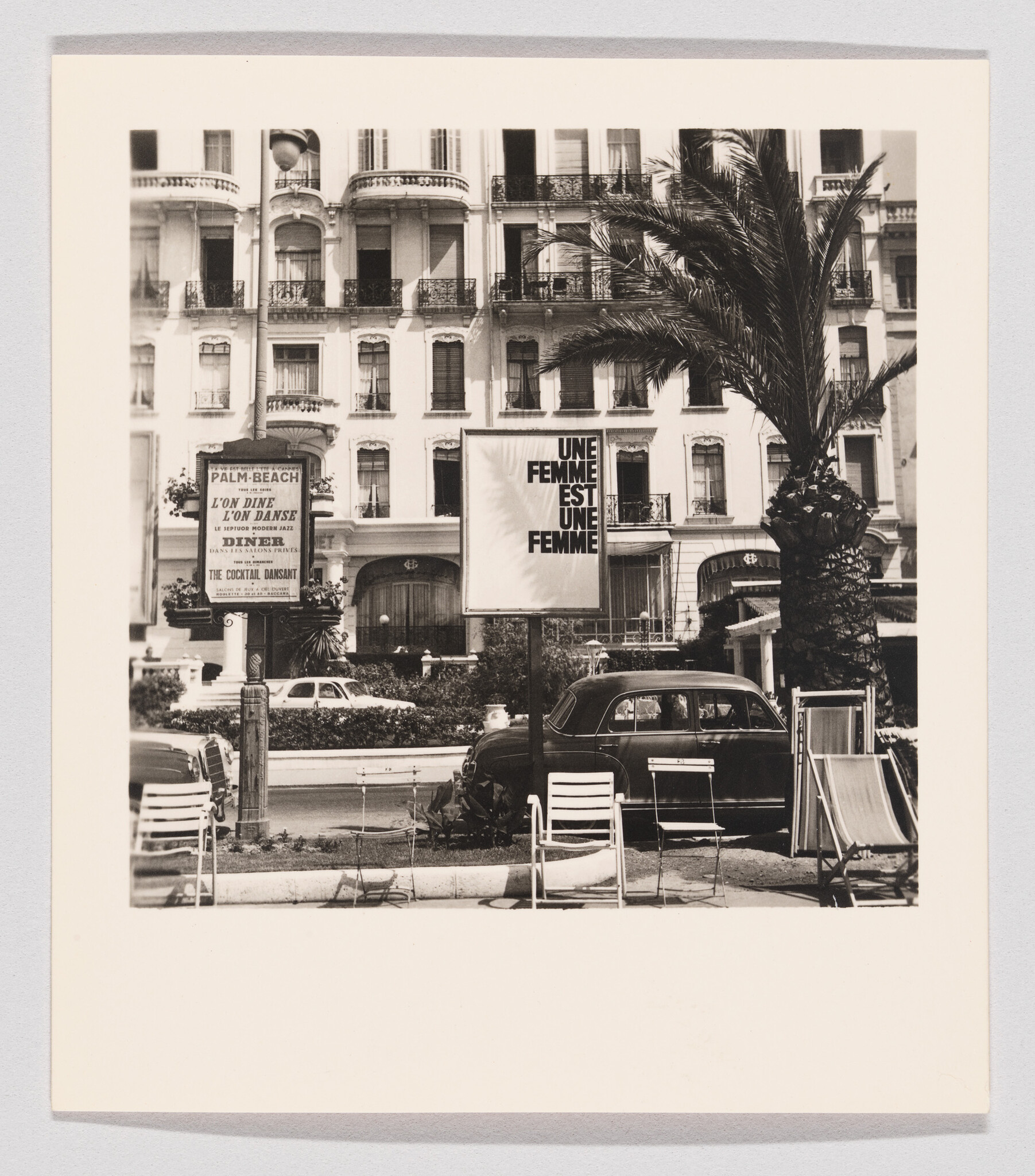 A vintage car parked by a palm tree under a billboard reading "Une femme est une femme."