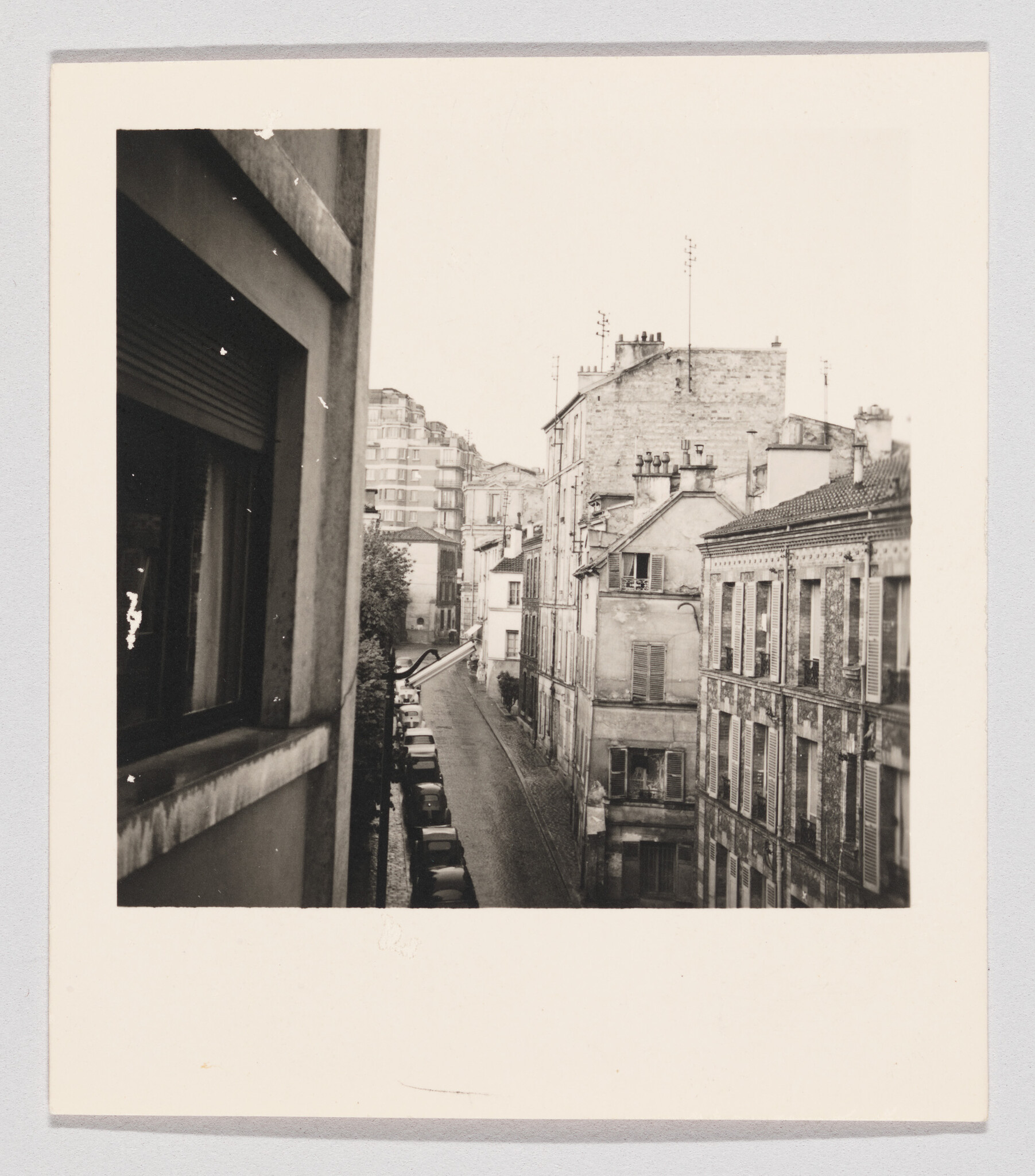 A narrow cobblestone street lined with parked cars and old apartment buildings seen from a window.