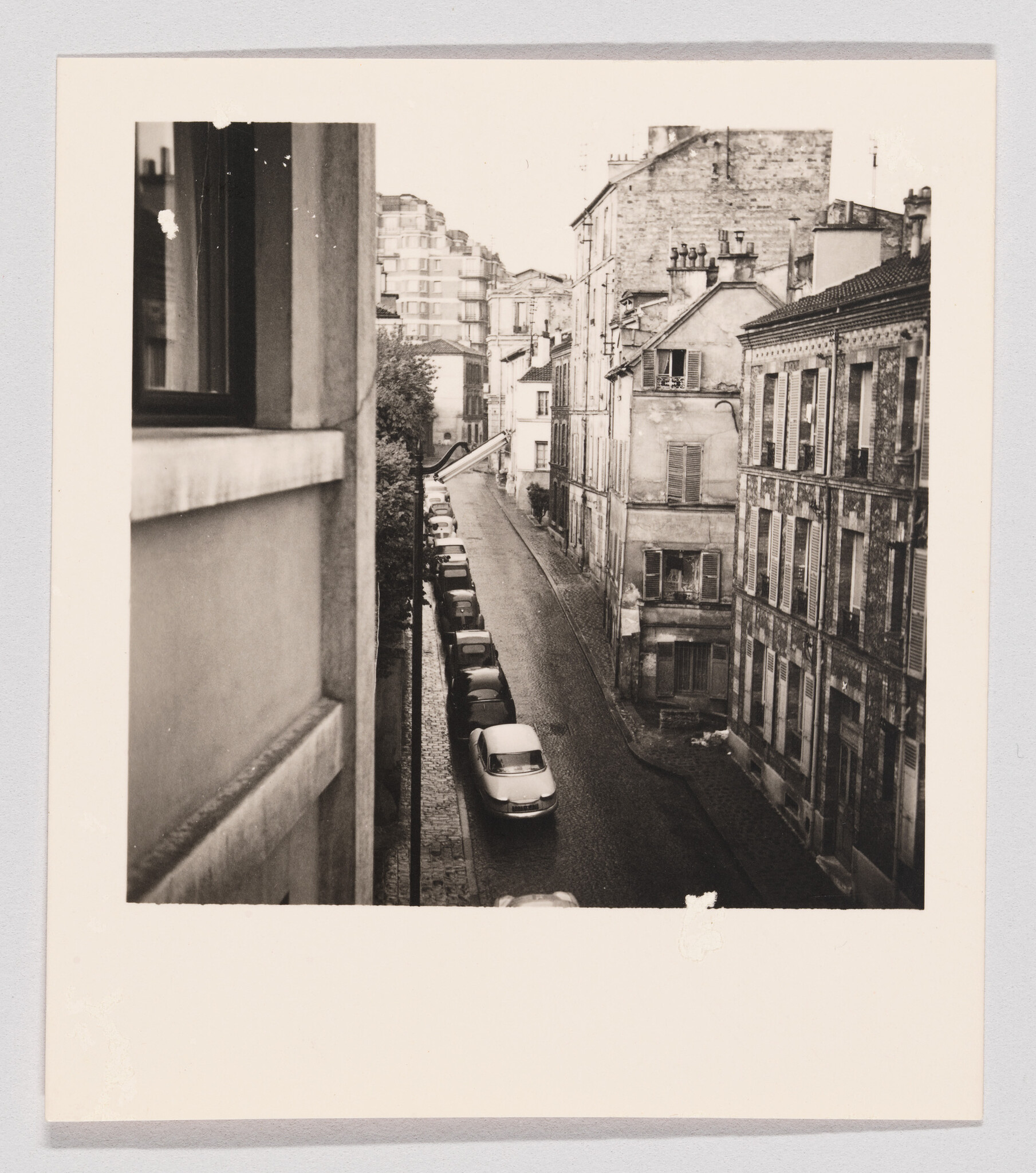 Narrow cobblestone street lined with parked cars between tall old apartment buildings.