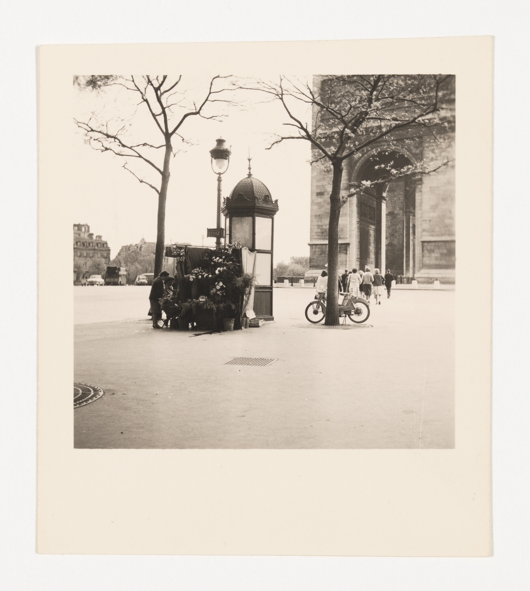 A street vendor arranges flowers beside a small kiosk while pedestrians walk toward a large stone arch.